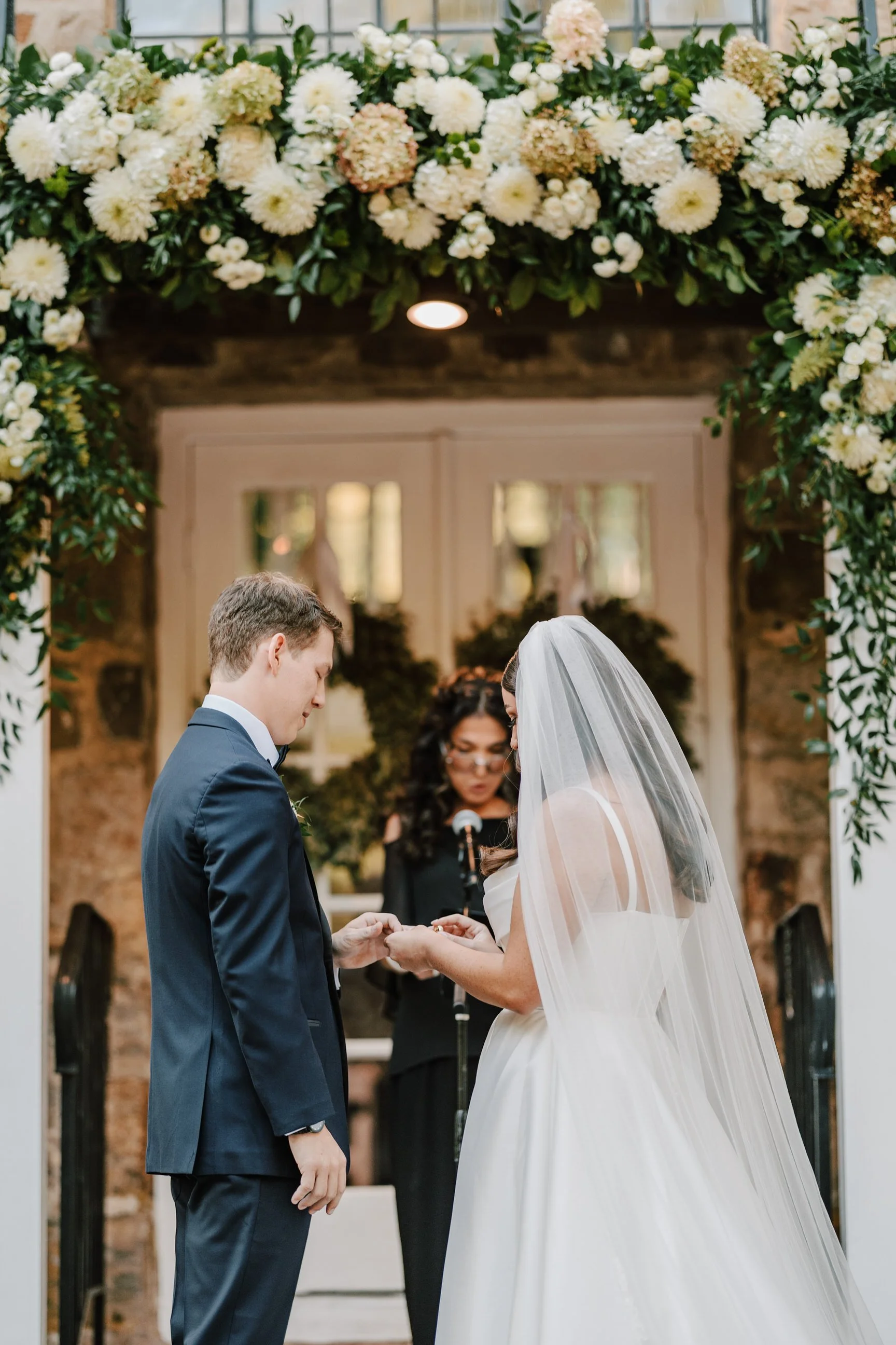 A bride and groom exchanging rings under a floral arch during a wedding ceremony indoors.