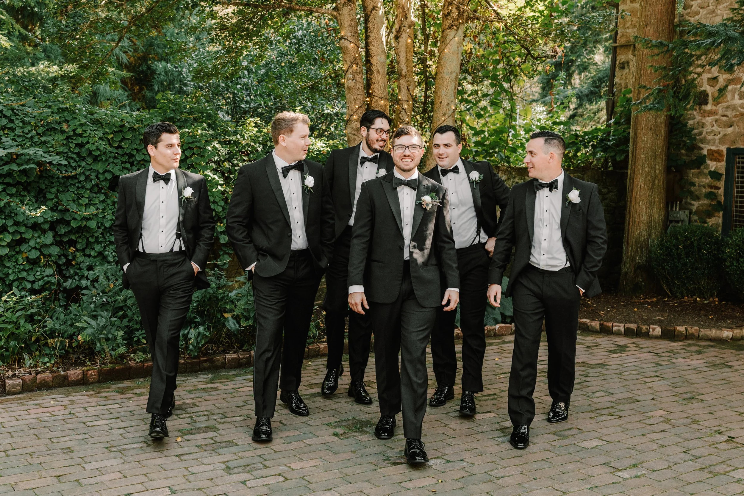 Group of seven men dressed in black tuxedos with bow ties, walking outdoors on a brick path surrounded by green trees and bushes, with sunlight filtering through the leaves.