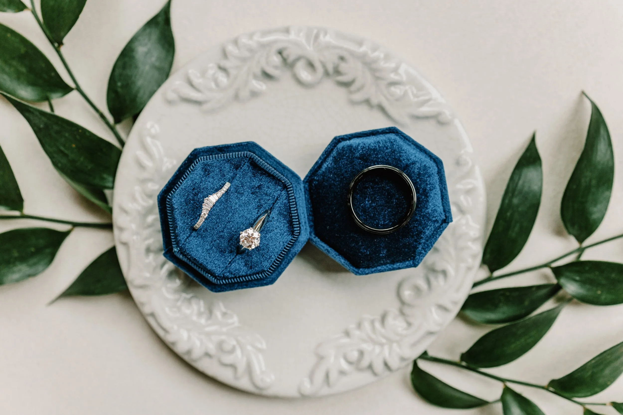 Open blue velvet ring box with a diamond engagement ring, along with a matching band, on a white decorative plate surrounded by green leaves.