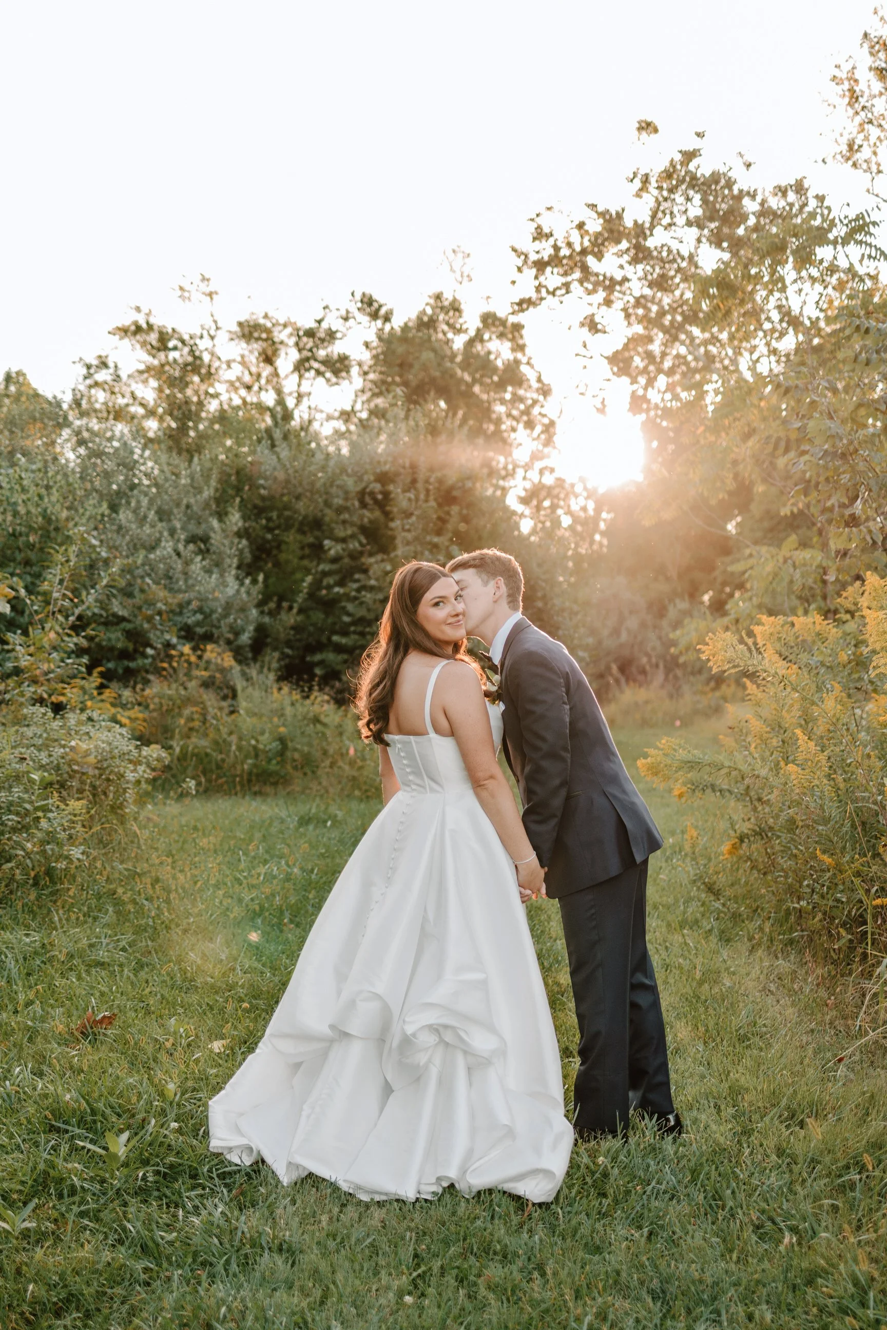 A bride and groom holding hands and sharing a kiss outdoors during sunset, with trees and greenery in the background.