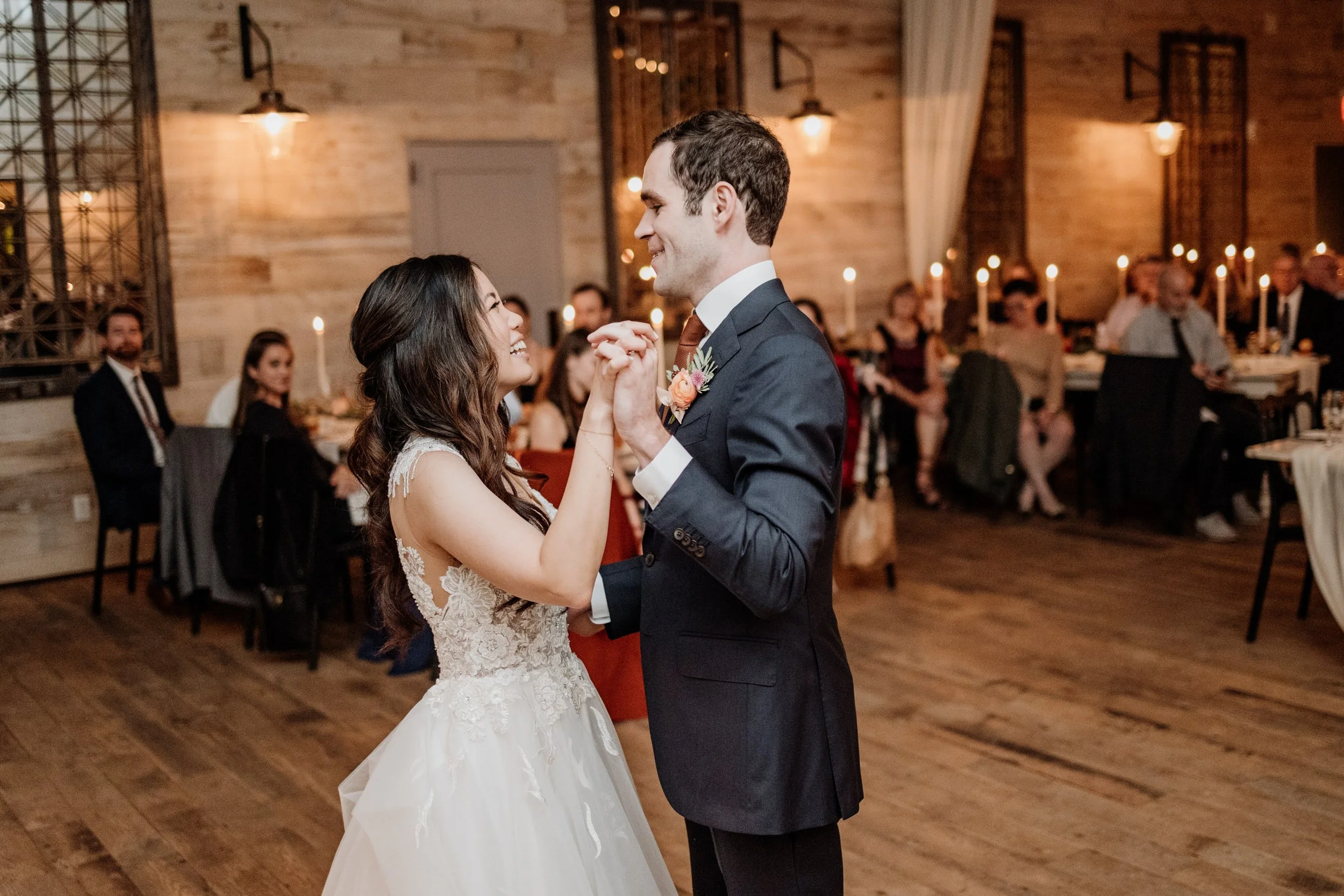 A bride and groom dancing at a wedding reception, smiling and holding hands, in a rustic decorated venue with guests seated in the background.