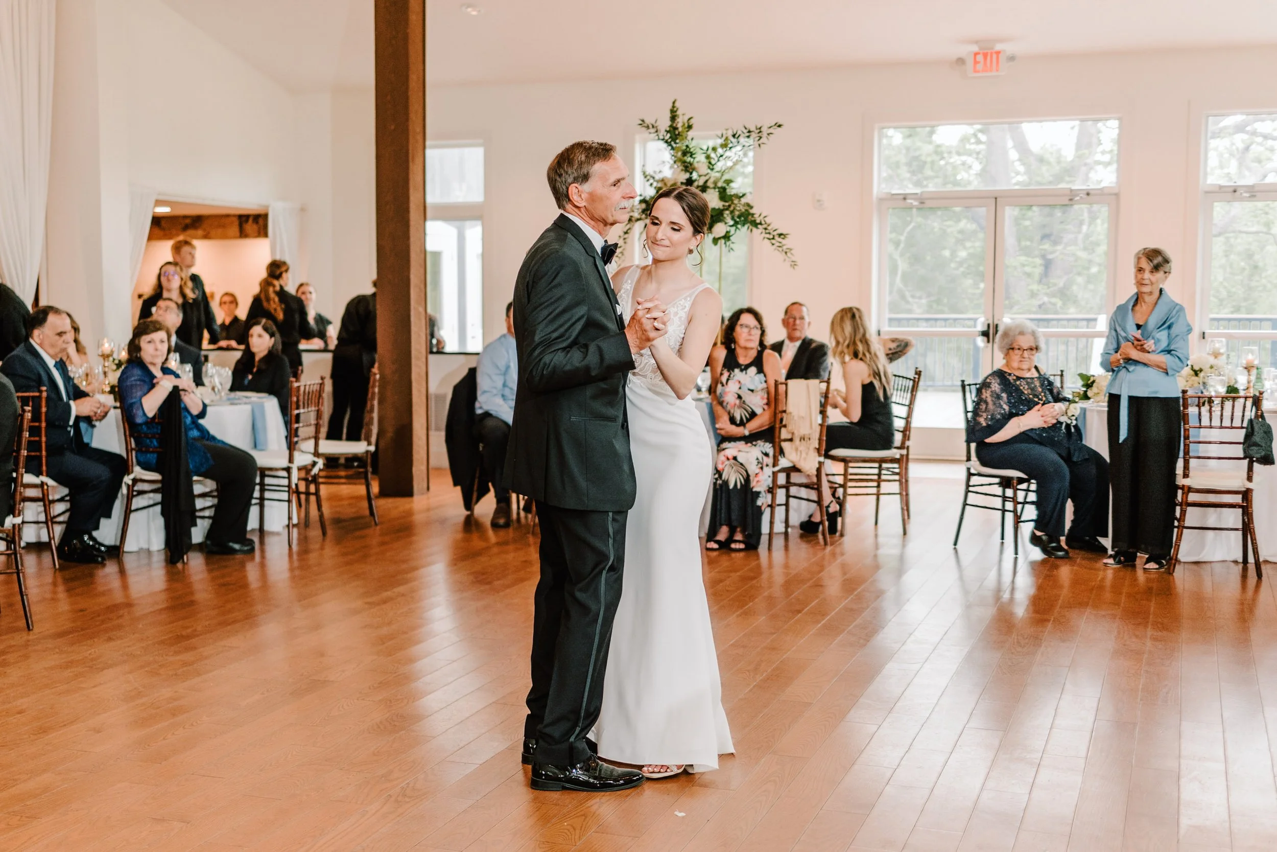 A bride and groom dancing during their wedding reception surrounded by seated guests in a brightly lit hall.