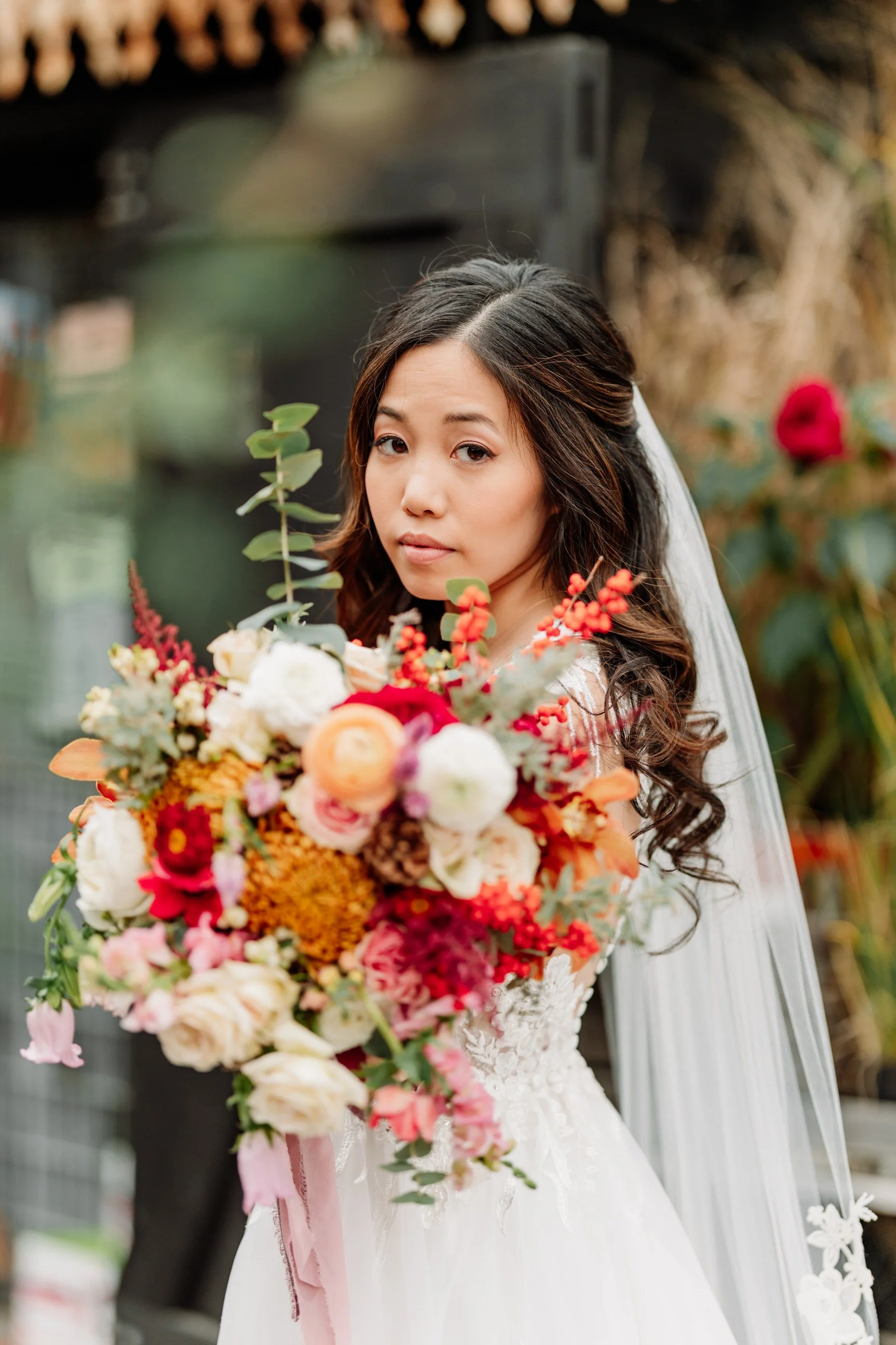 A bride holding a large colorful bouquet of flowers, looking over her shoulder, outdoors with greenery and flowers in the background.