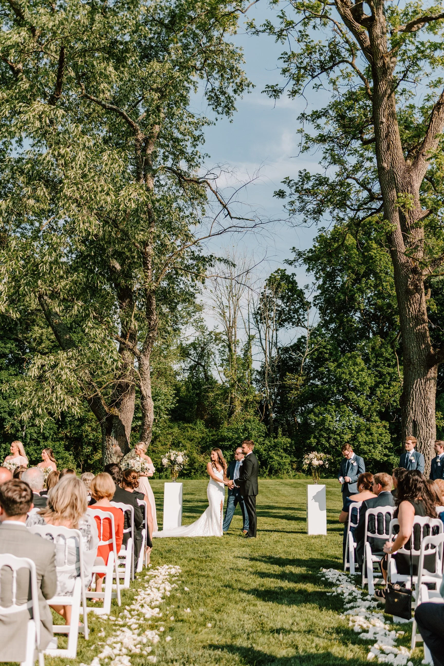 Outdoor wedding ceremony with a bride and groom exchanging vows, surrounded by seated guests, officiant, bridesmaids, and groomsmen, set in a lush green park with tall trees and white flower petals on the grass.