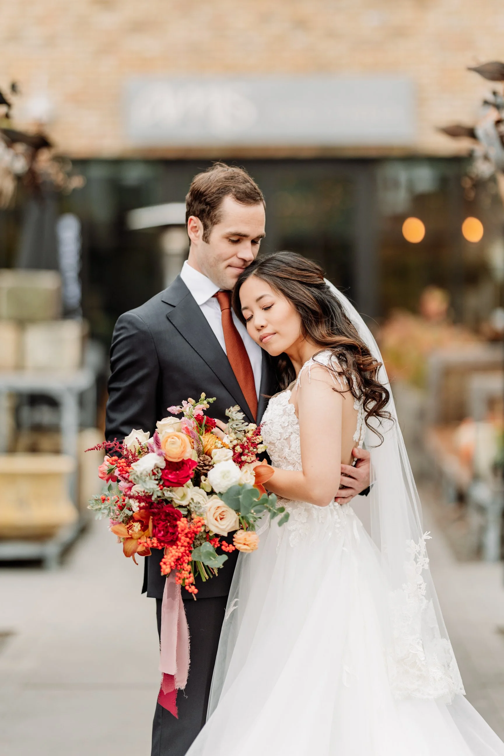 A bride and groom embrace outdoors, the bride holding a vibrant bouquet of flowers, with a blurred background of a building and warm lighting.