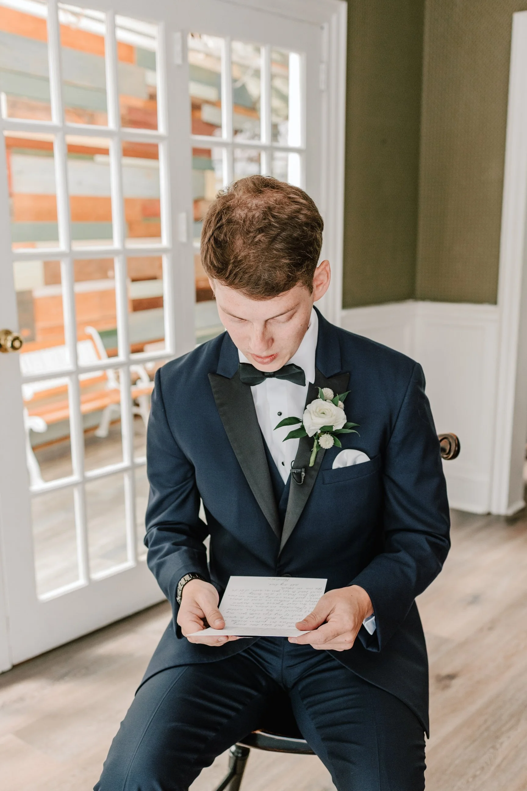 A young man in a tuxedo reading a note or letter in a bright room with large porcelain doors and wooden flooring.