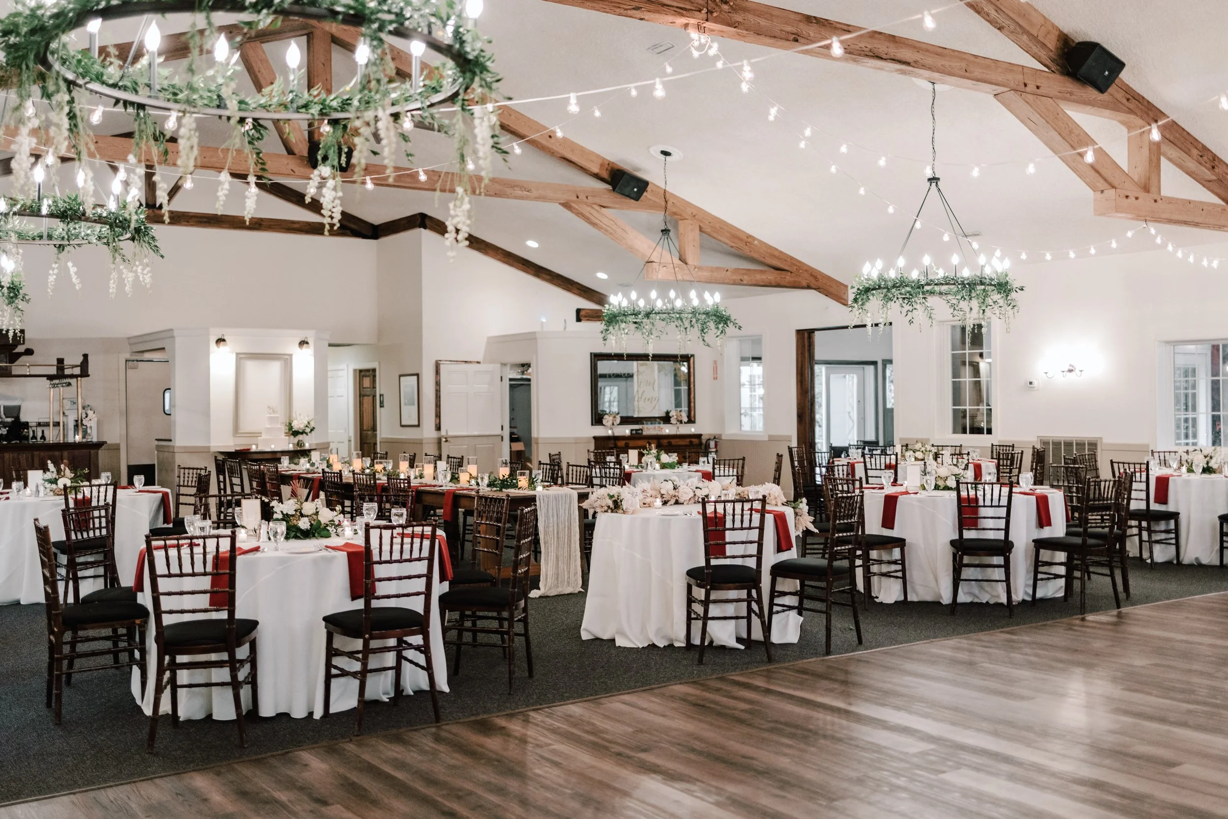 A banquet hall decorated with string lights and hanging greenery chandeliers, featuring round tables with white tablecloths, red napkins, and floral centerpieces, set up for a formal event.