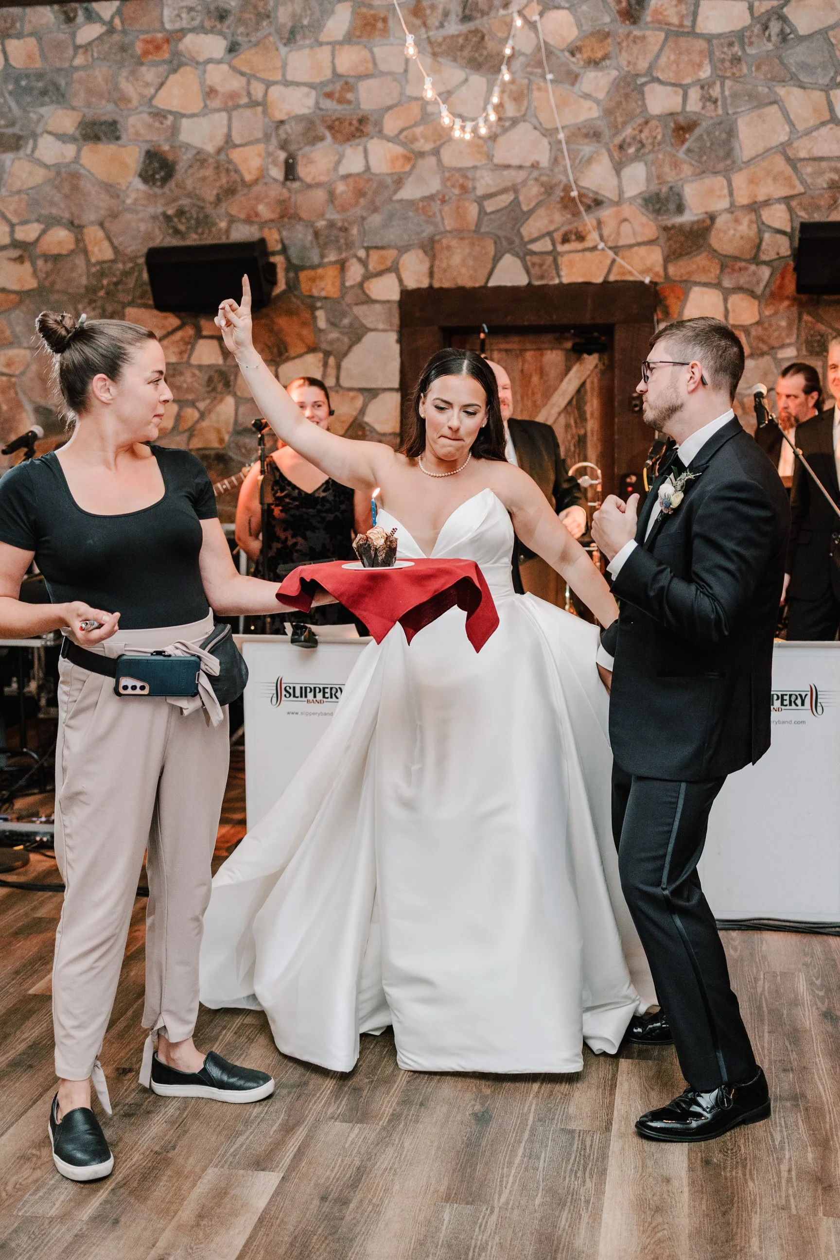 Wedding reception with bride and groom humorously dancing, bride holding a cupcake on a red napkin, woman in black shirt and beige pants watching, band in the background, stone wall decor.