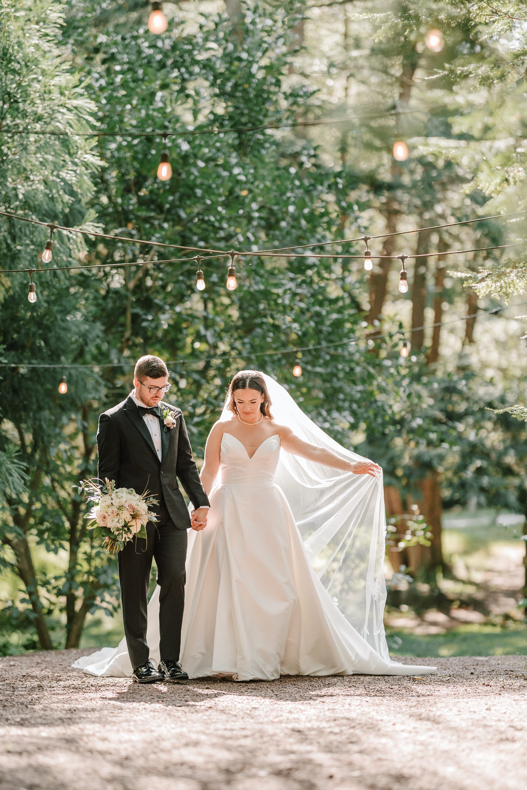 Bride and groom holding hands in a wooded outdoor setting, with string lights overhead. The bride is in a white wedding gown holding the train, and the groom is in a black tuxedo holding a bouquet of flowers.