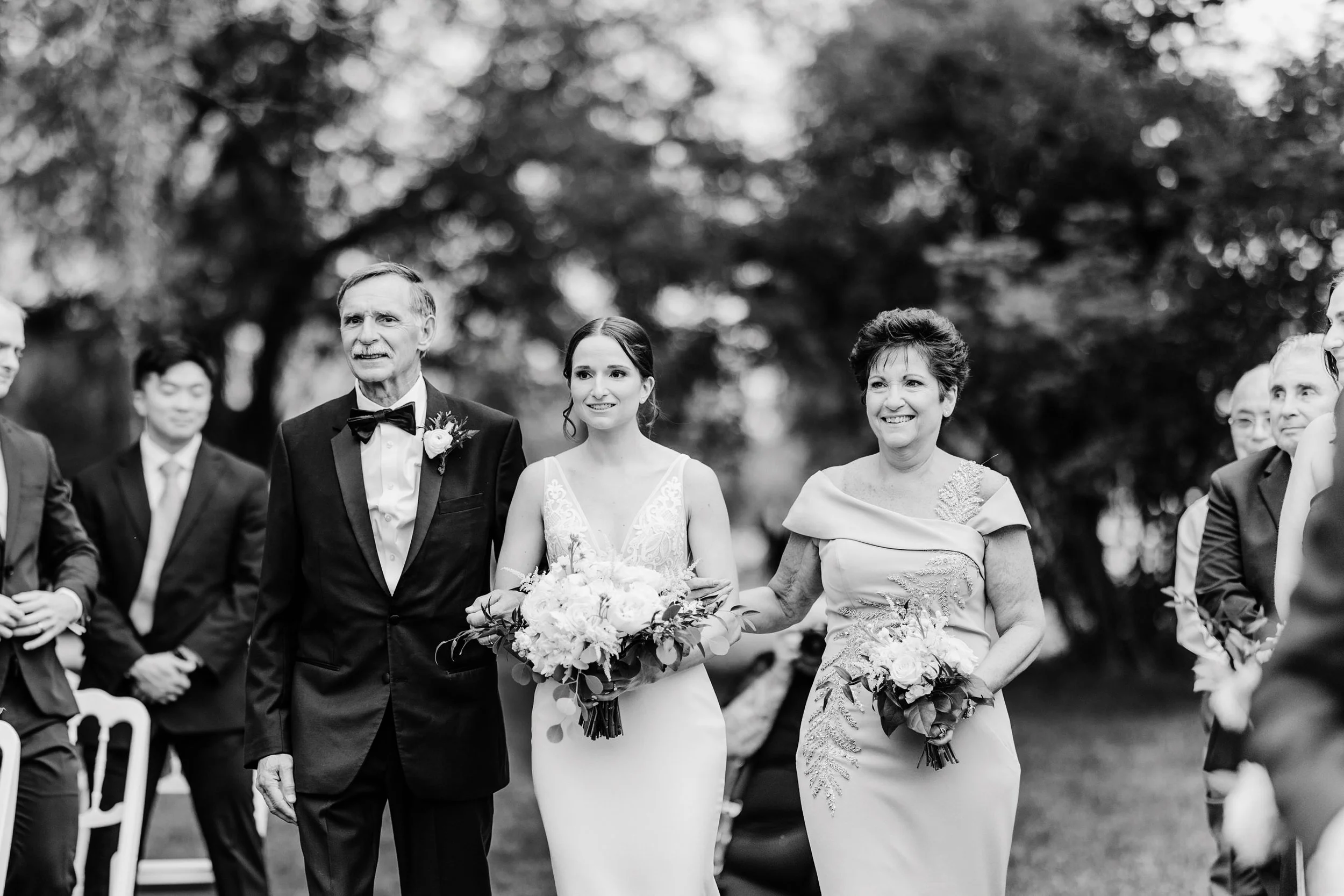 Black and white photo of a wedding ceremony outdoors, with a woman in a wedding dress holding a bouquet, escorted by a man in a tuxedo and a woman in an elegant dress carrying a bouquet. Other guests are seated and standing in the background.