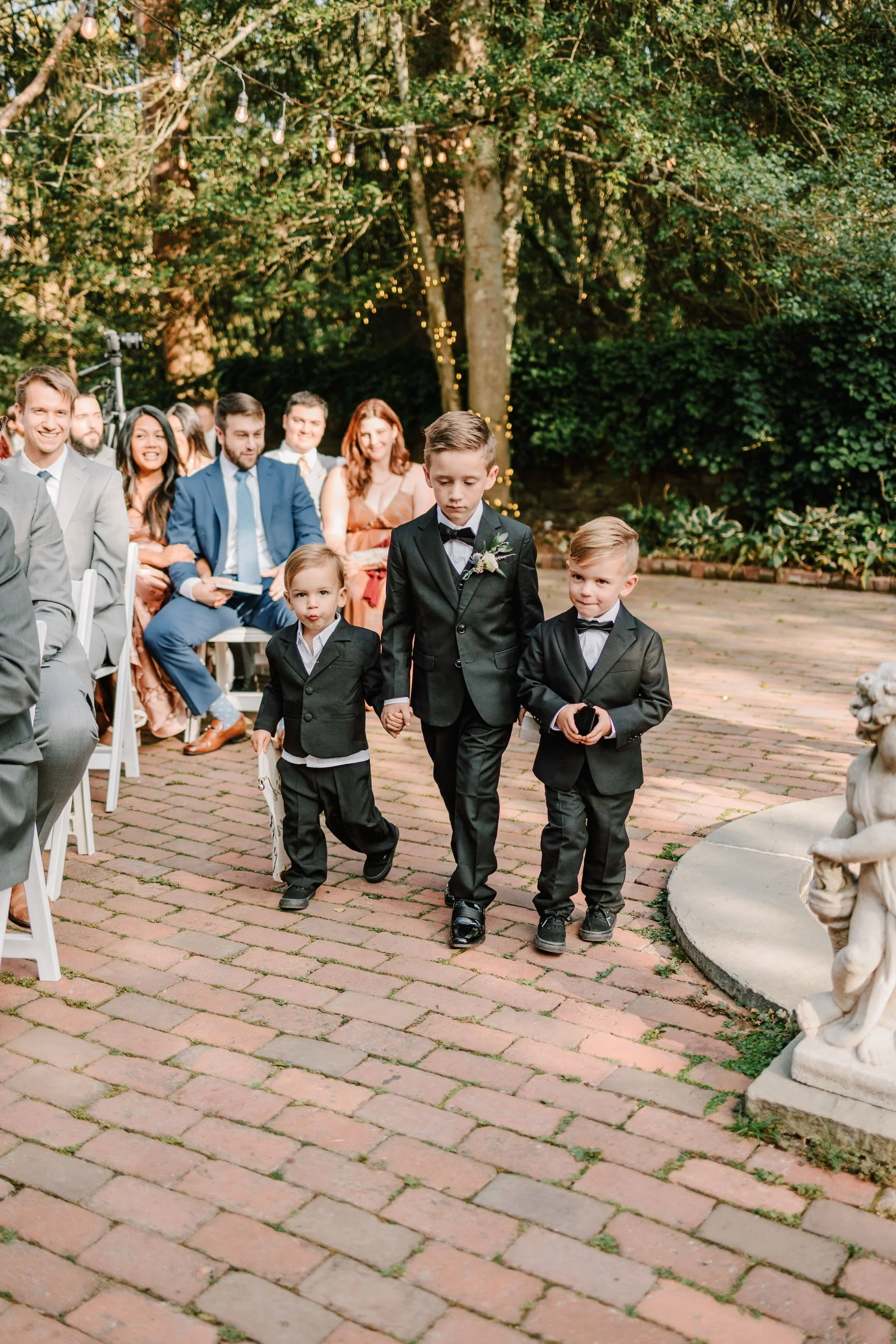 Three young boys dressed in suits walk down an outdoor brick pathway during a wedding ceremony, with seated guests watching.