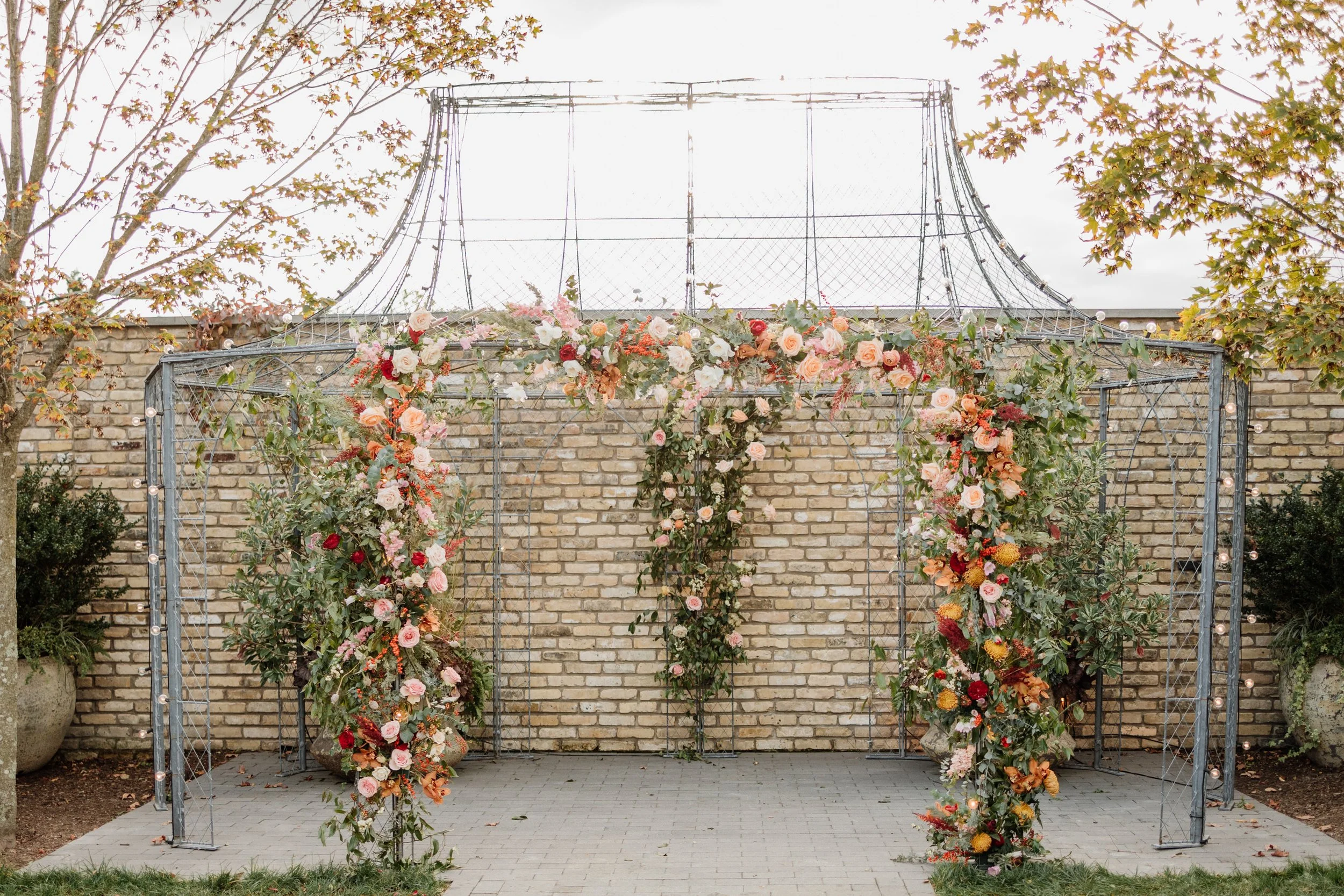 Outdoor wedding or event arch decorated with pink, orange, and white flowers, greenery, and string lights, set against a brick wall on a paved area.