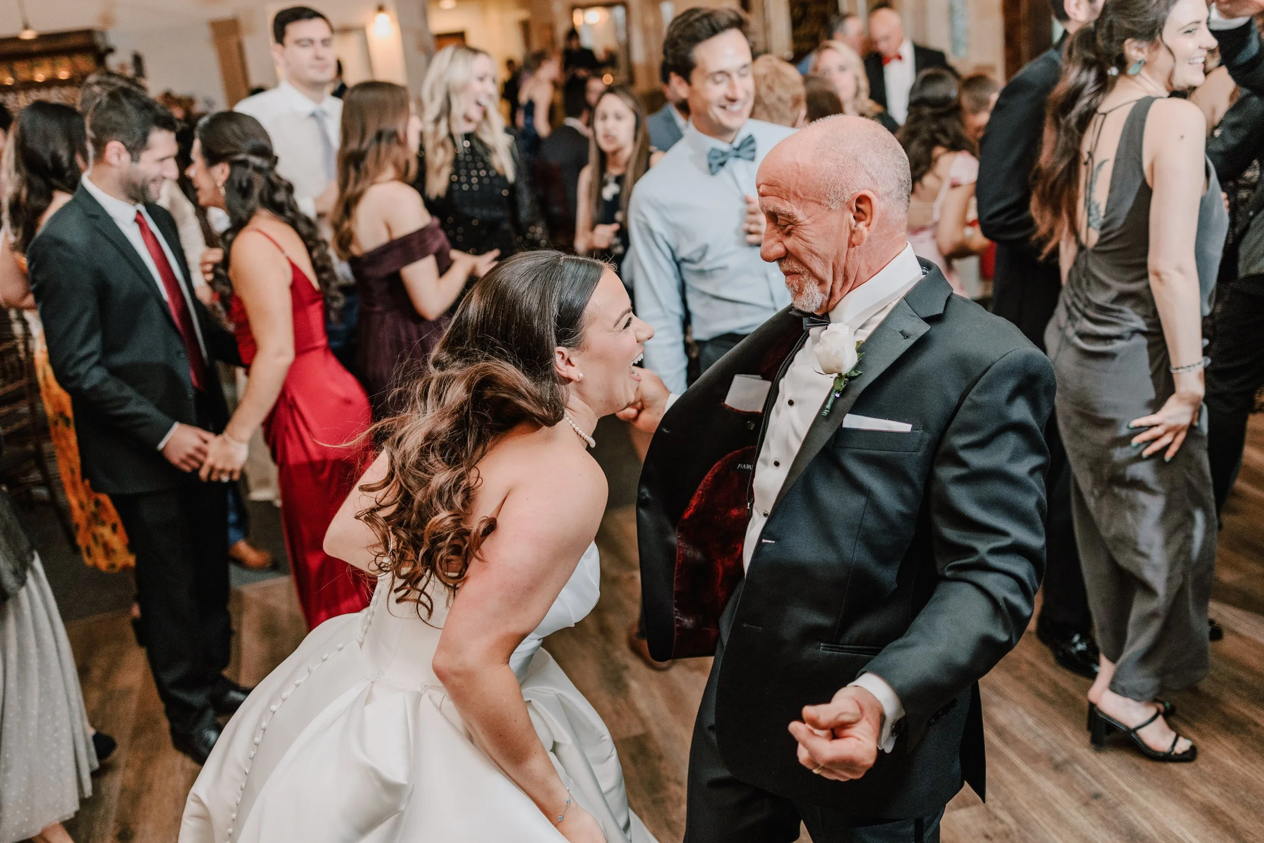 A bride and an older man dancing and smiling at a wedding reception with other guests dancing and socializing in the background.