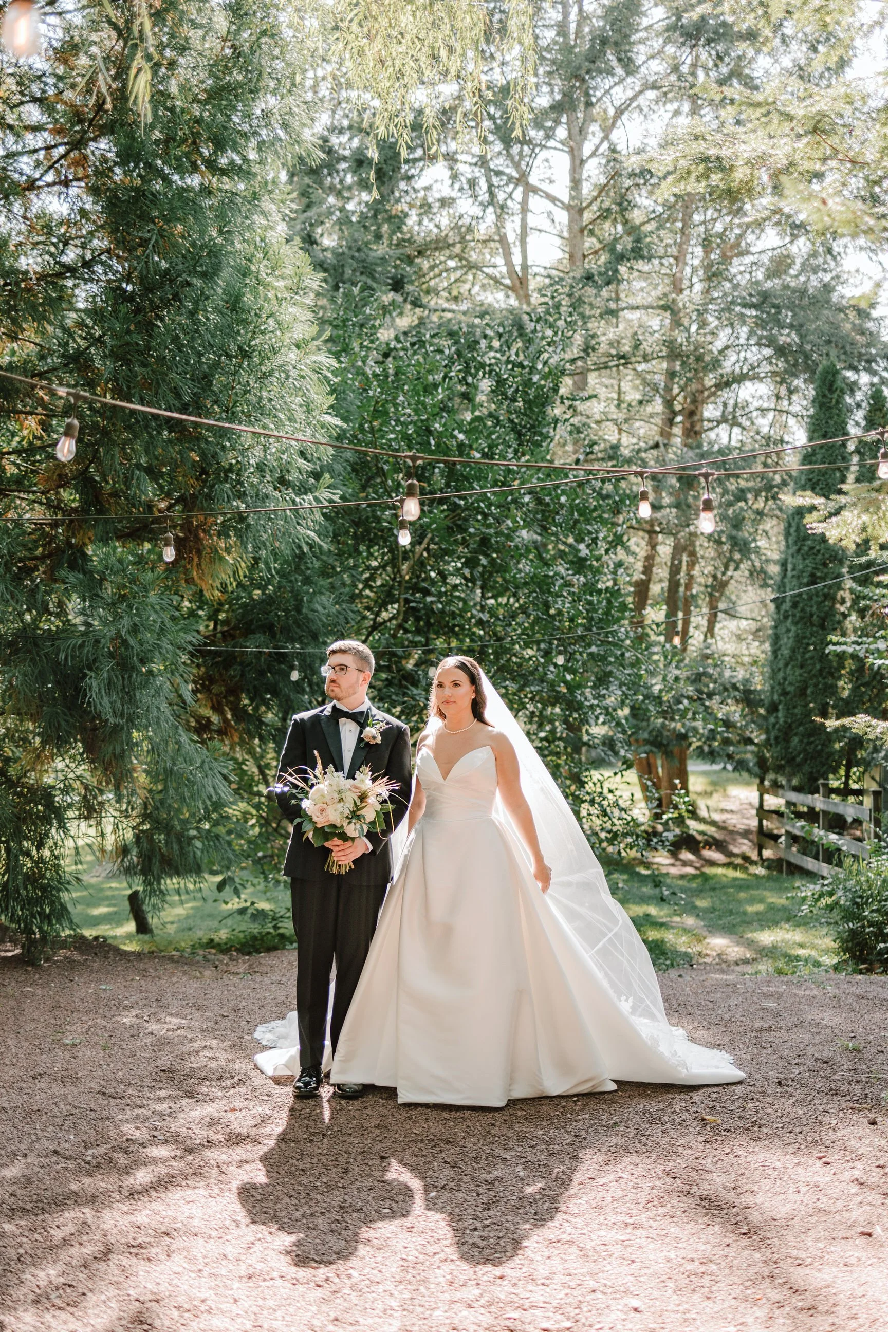 Bride and groom standing outdoors under string lights, trees and greenery in background, the bride in a white wedding dress holding onto the groom, who is dressed in a black tuxedo holding a bouquet of flowers.