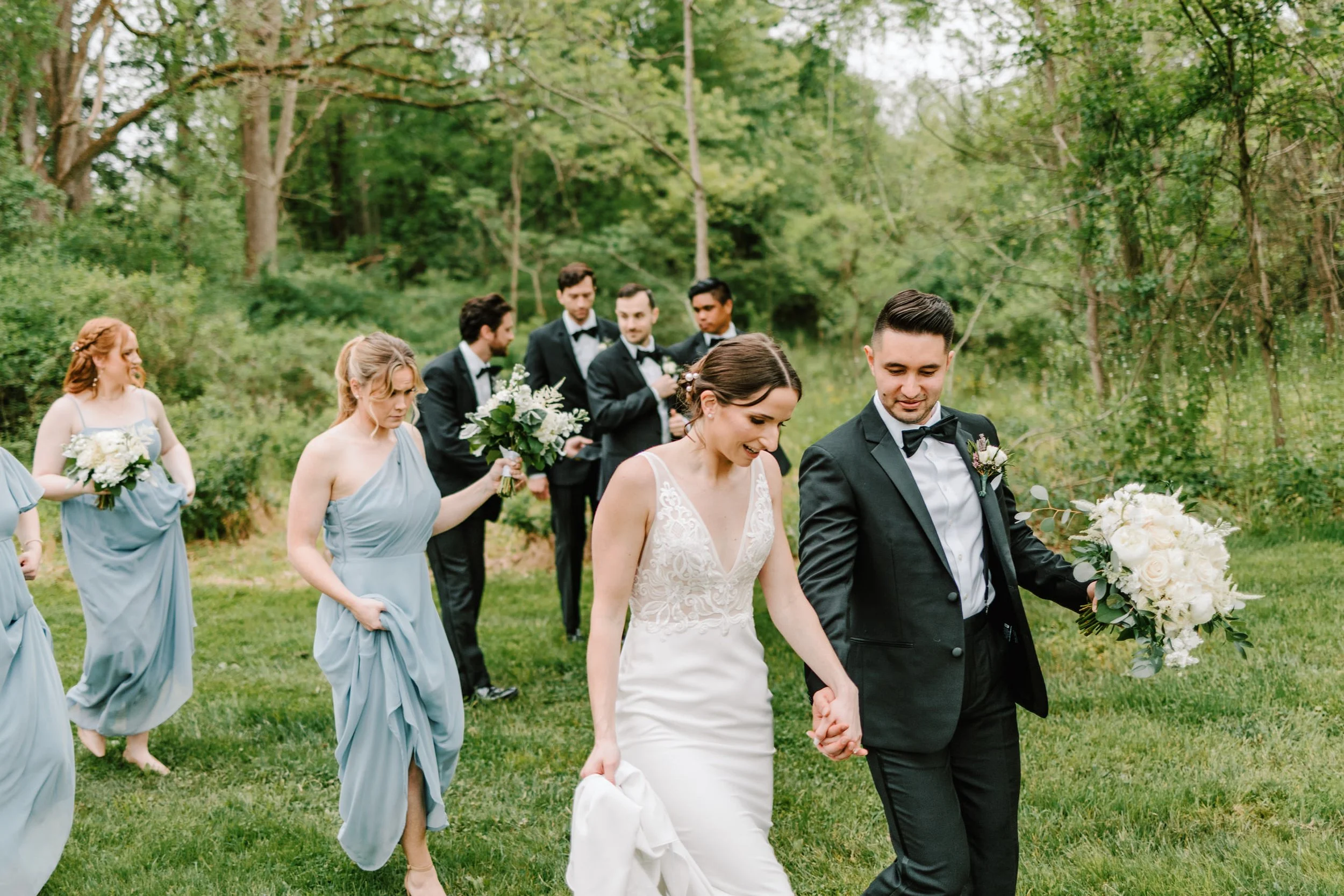 A bride and groom walk hand in hand through a grassy outdoor area, surrounded by bridesmaids and groomsmen, during a wedding ceremony. The bride is wearing a white lace dress, and the groom is in a black tuxedo with a bow tie. The bridesmaids are in 