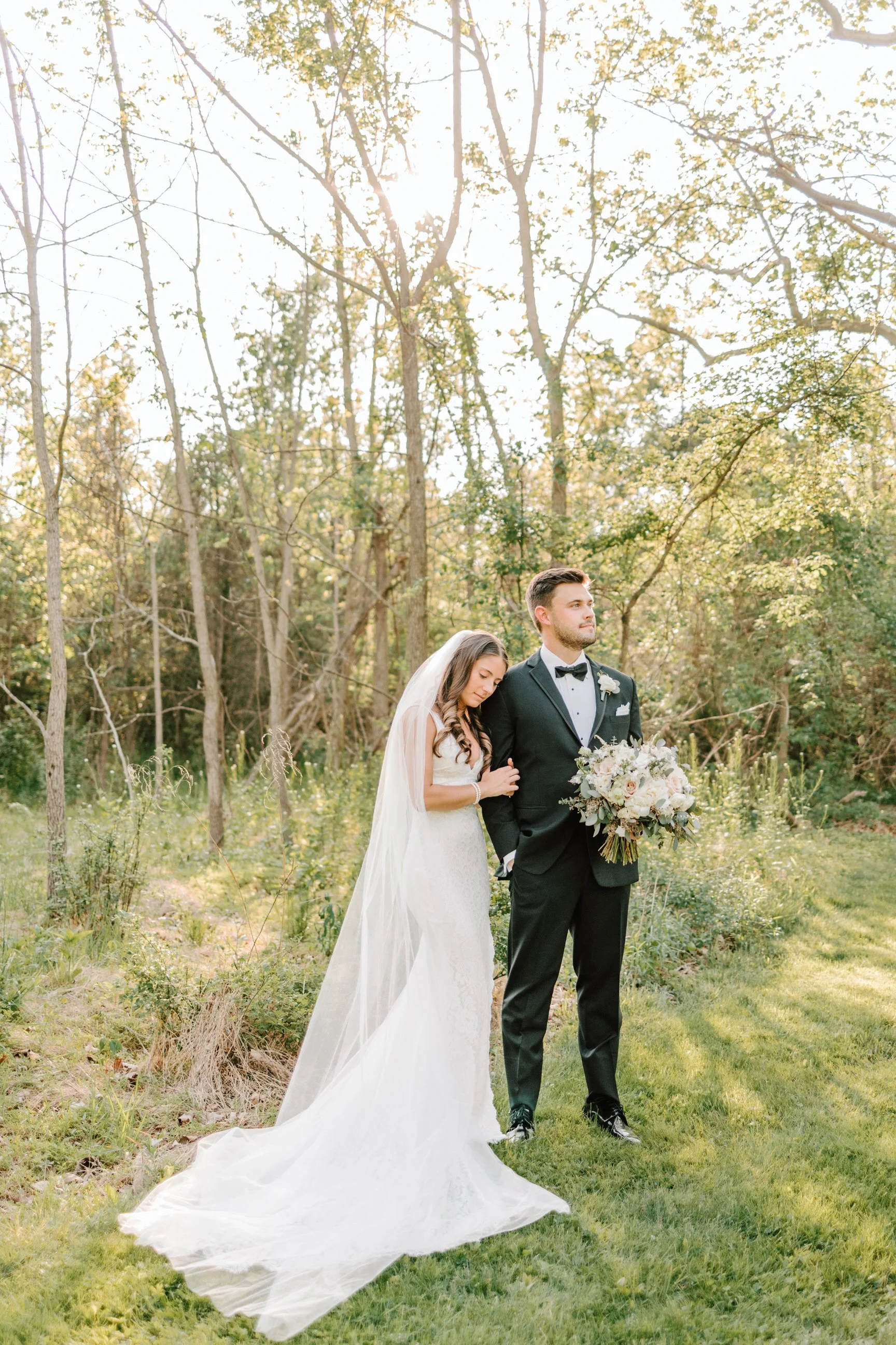 A bride and groom standing outdoors among trees during a wedding photoshoot, with sunlight filtering through the trees.