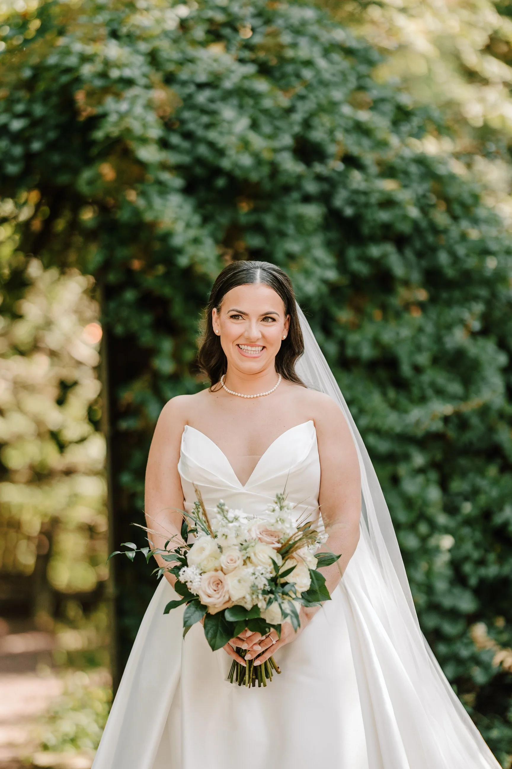 A bride in a white wedding dress holding a bouquet of white and blush roses and greenery, smiling outdoors with a green leafy background.