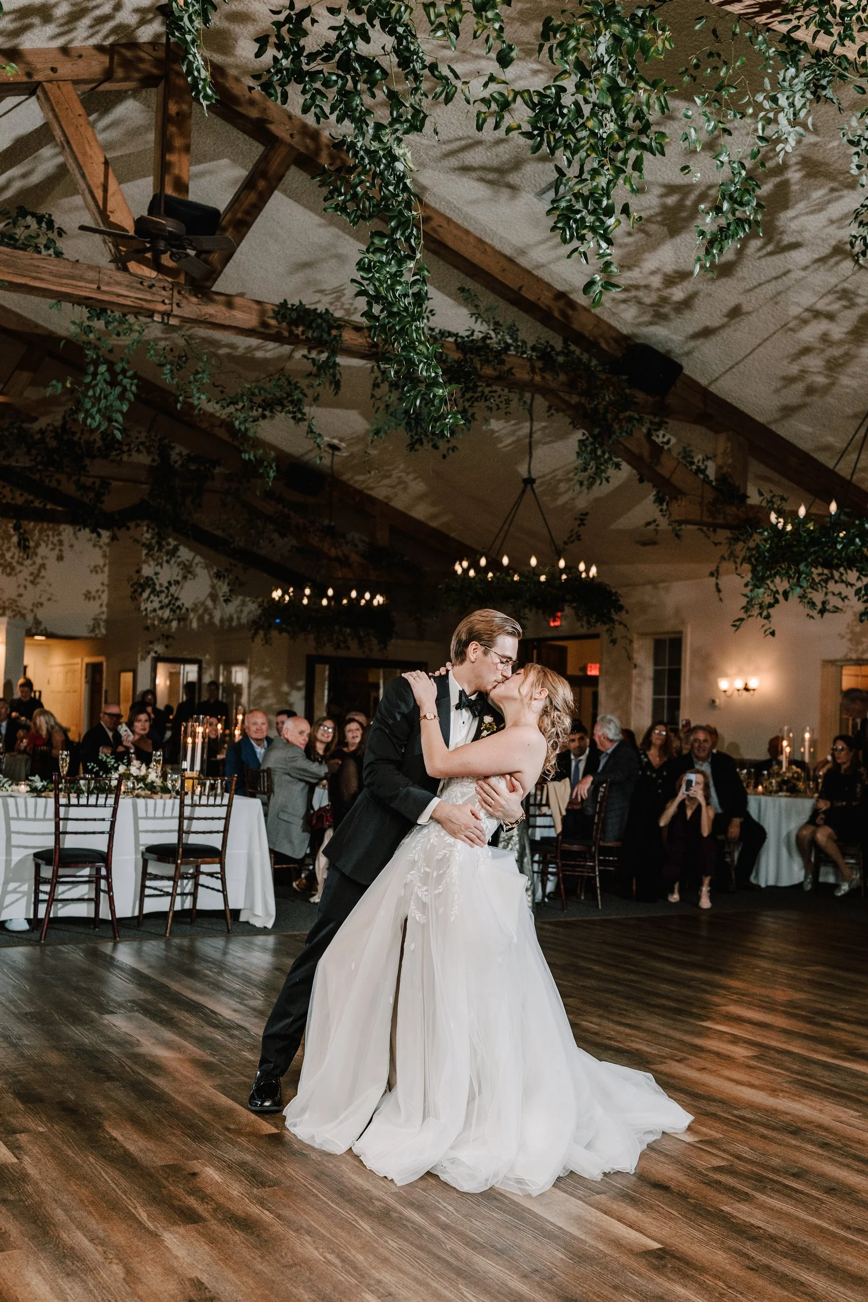 A newly married couple sharing a kiss on their wedding dance floor surrounded by seated guests at a wedding reception.