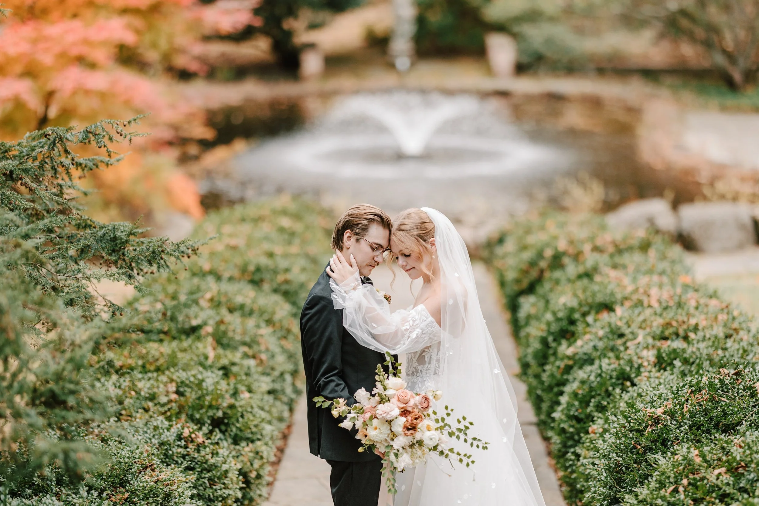 A bride and groom embrace each other on a pathway by a pond with a fountain, surrounded by autumn trees and bushes. Holly Hedge Estate Sharyn Louise Photo