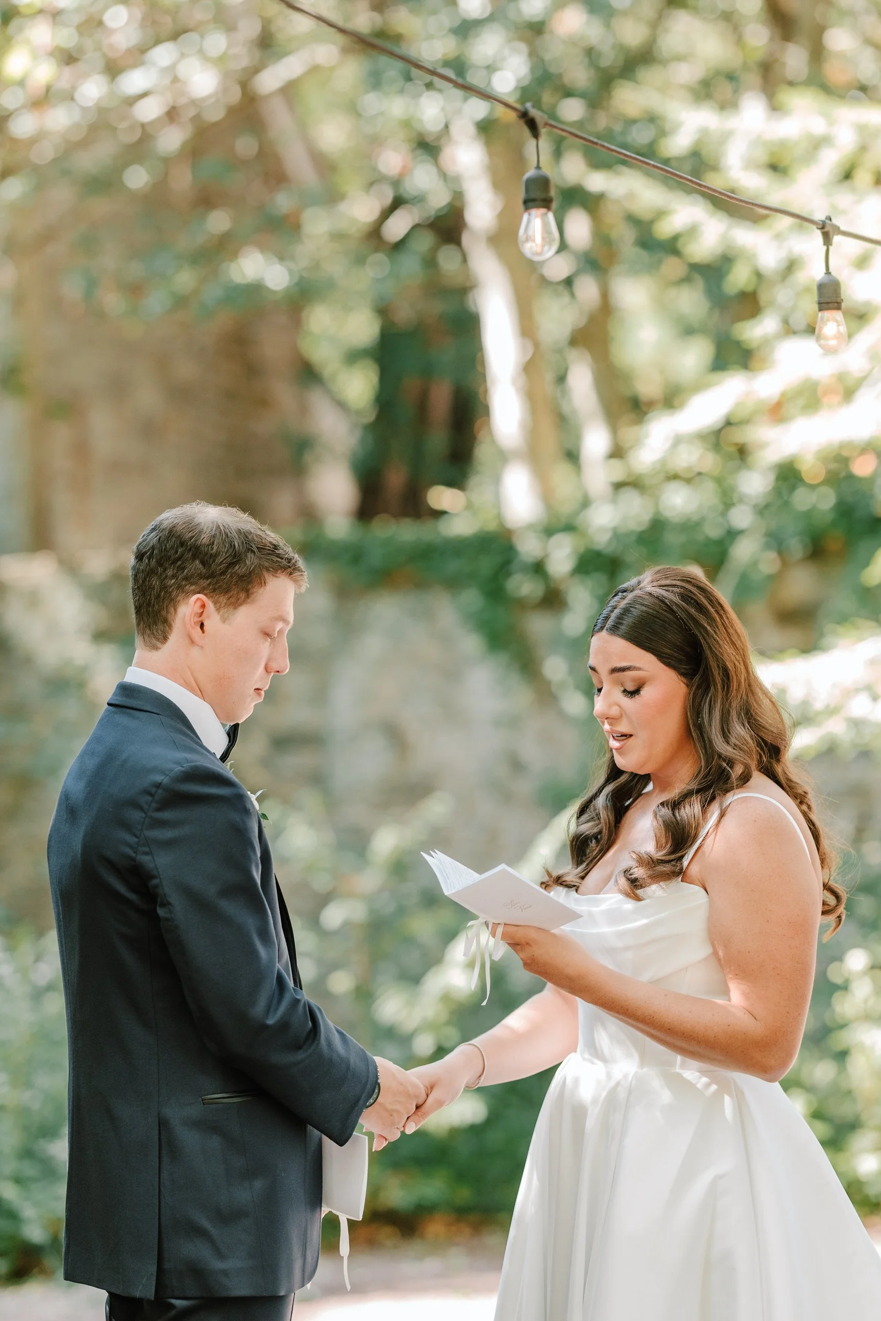 A bride and groom holding hands during a wedding ceremony outdoors, with the bride reading vows from a booklet, surrounded by greenery and string lights.