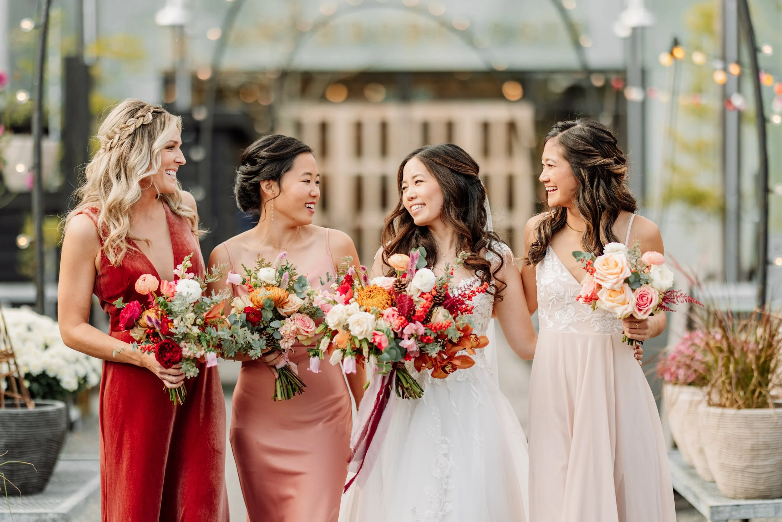 Five women in colorful dresses holding bouquets, smiling and looking at each other in a greenhouse or garden setting.