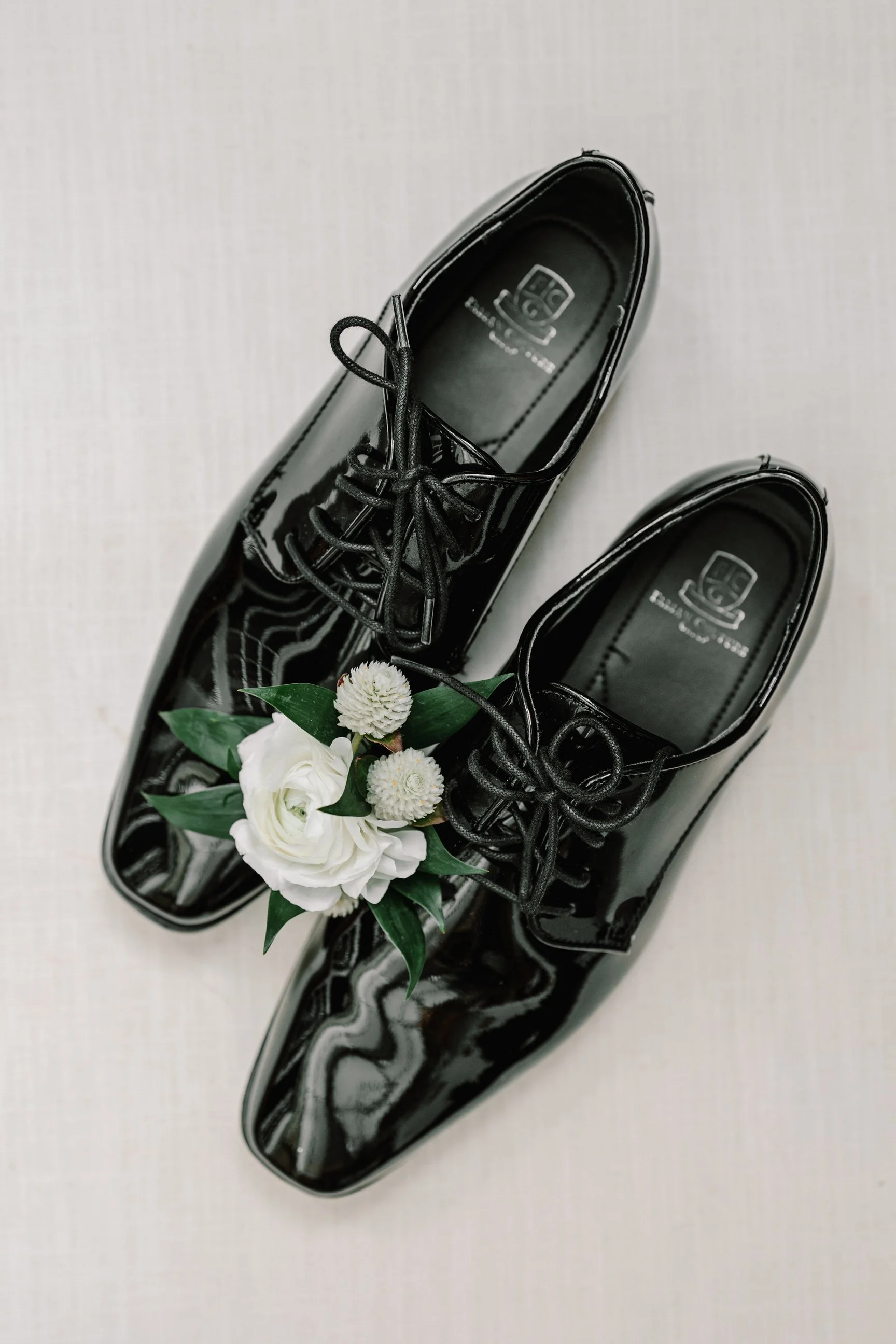 Black shiny dress shoes with a small bouquet of white flowers placed between them on a white surface.