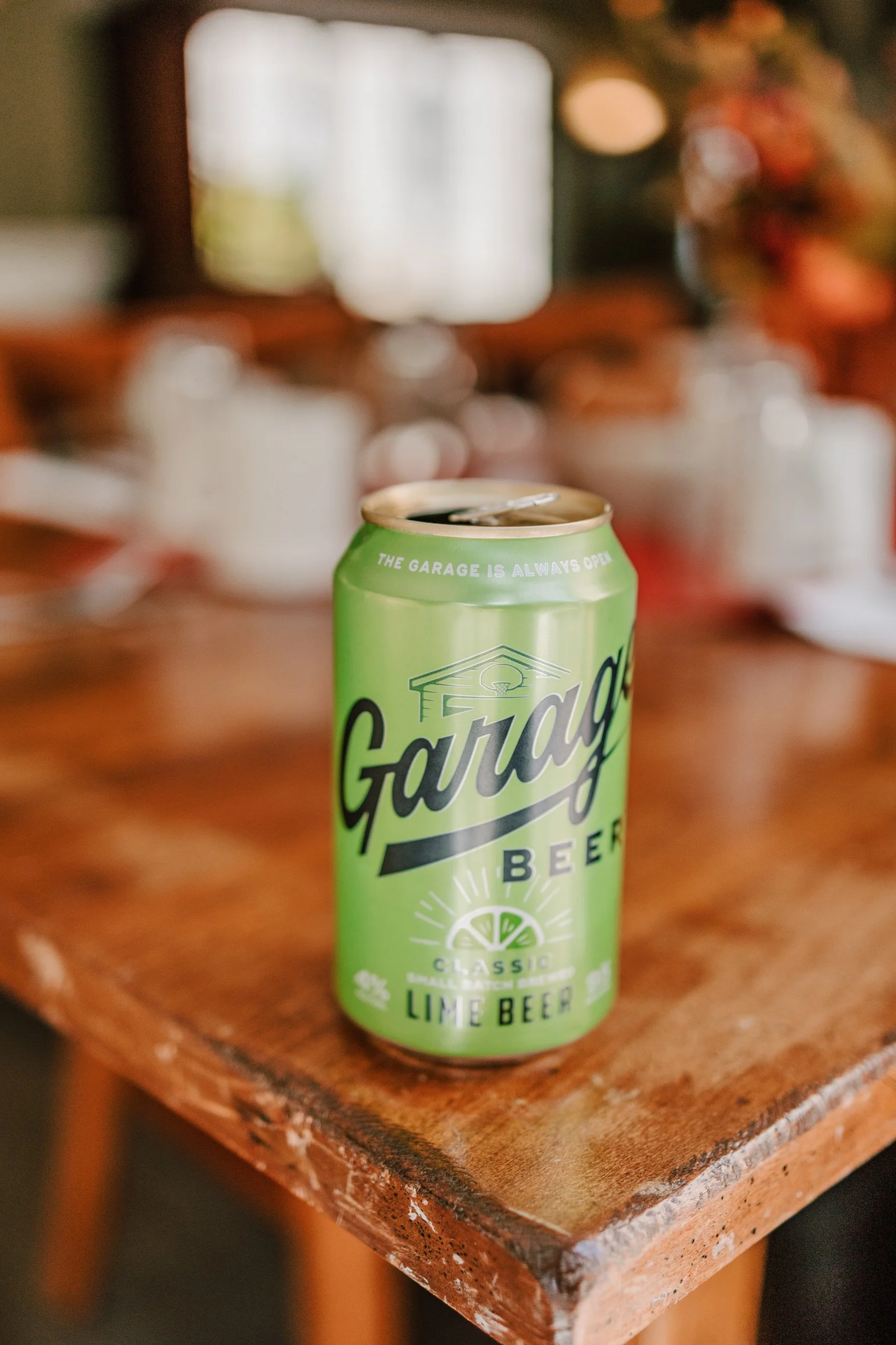 A green can of Garage Beer lime beer on a rustic wooden table with a blurred background of a window and various objects.
