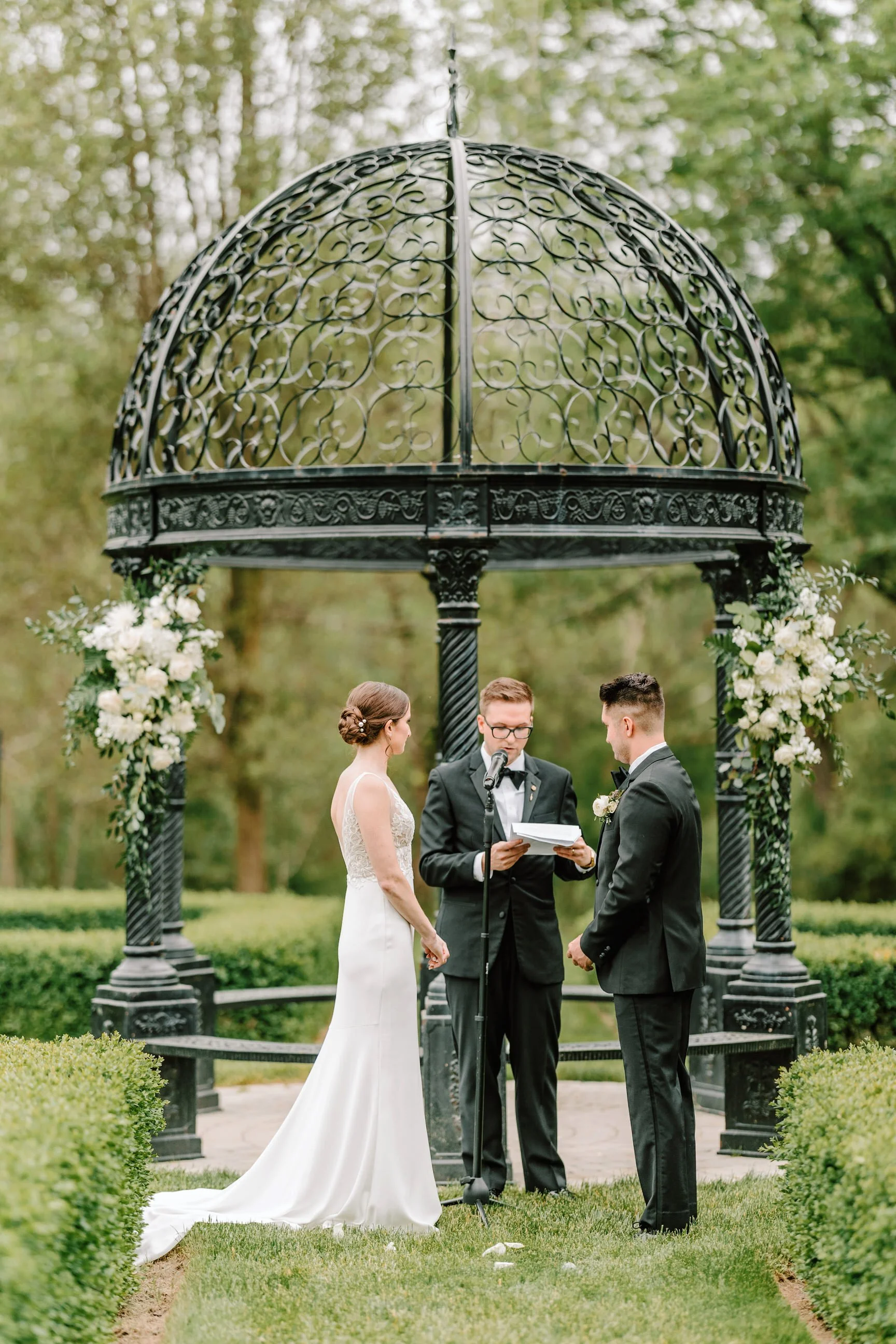 A wedding ceremony taking place outdoors under a decorative black wrought-iron gazebo adorned with white flowers, with a bride and groom standing before an officiant, surrounded by greenery.