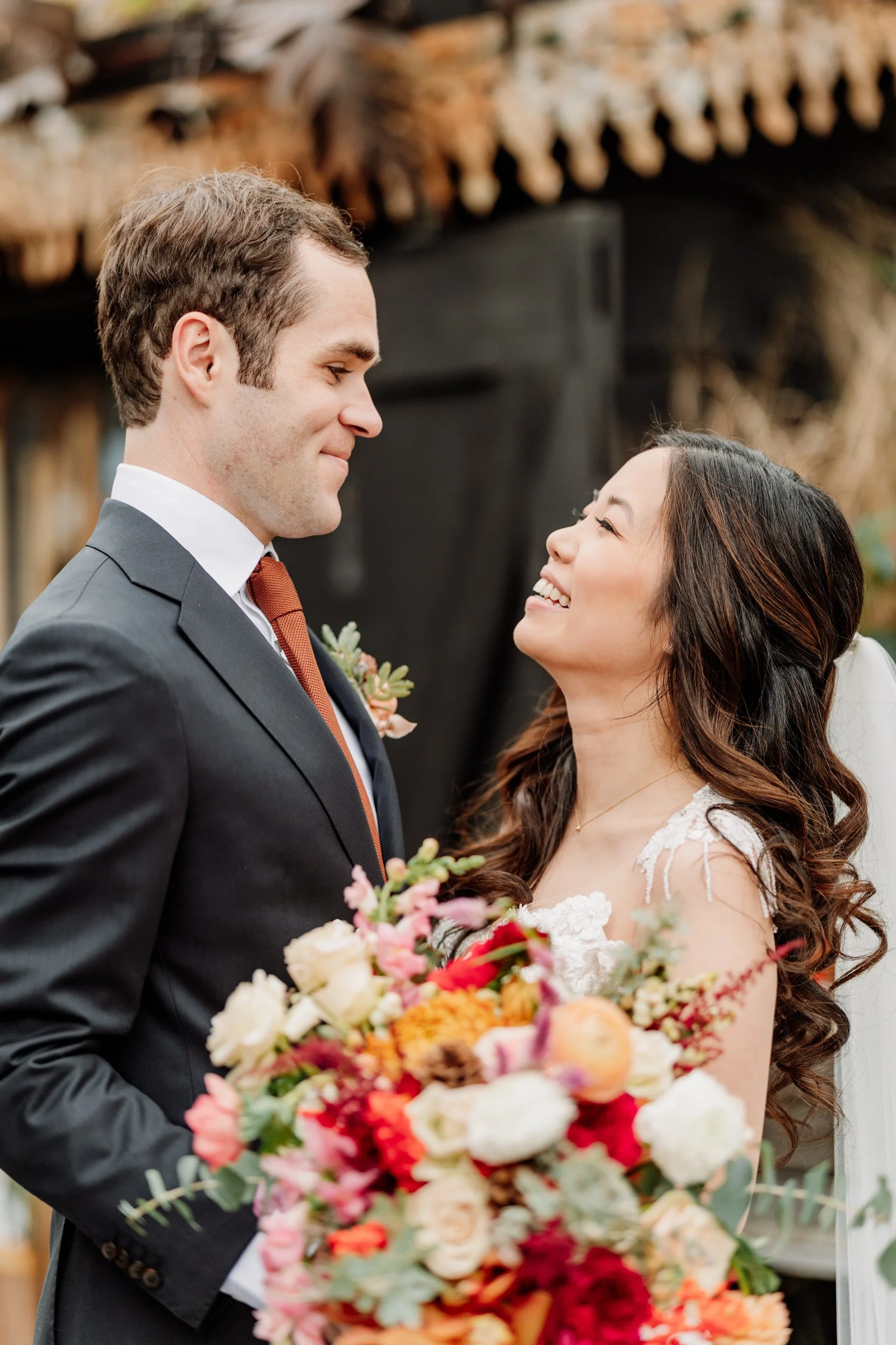 A bride and groom sharing a tender moment during their wedding, with the bride holding a large colorful bouquet of flowers, standing outdoors with a rustic backdrop.