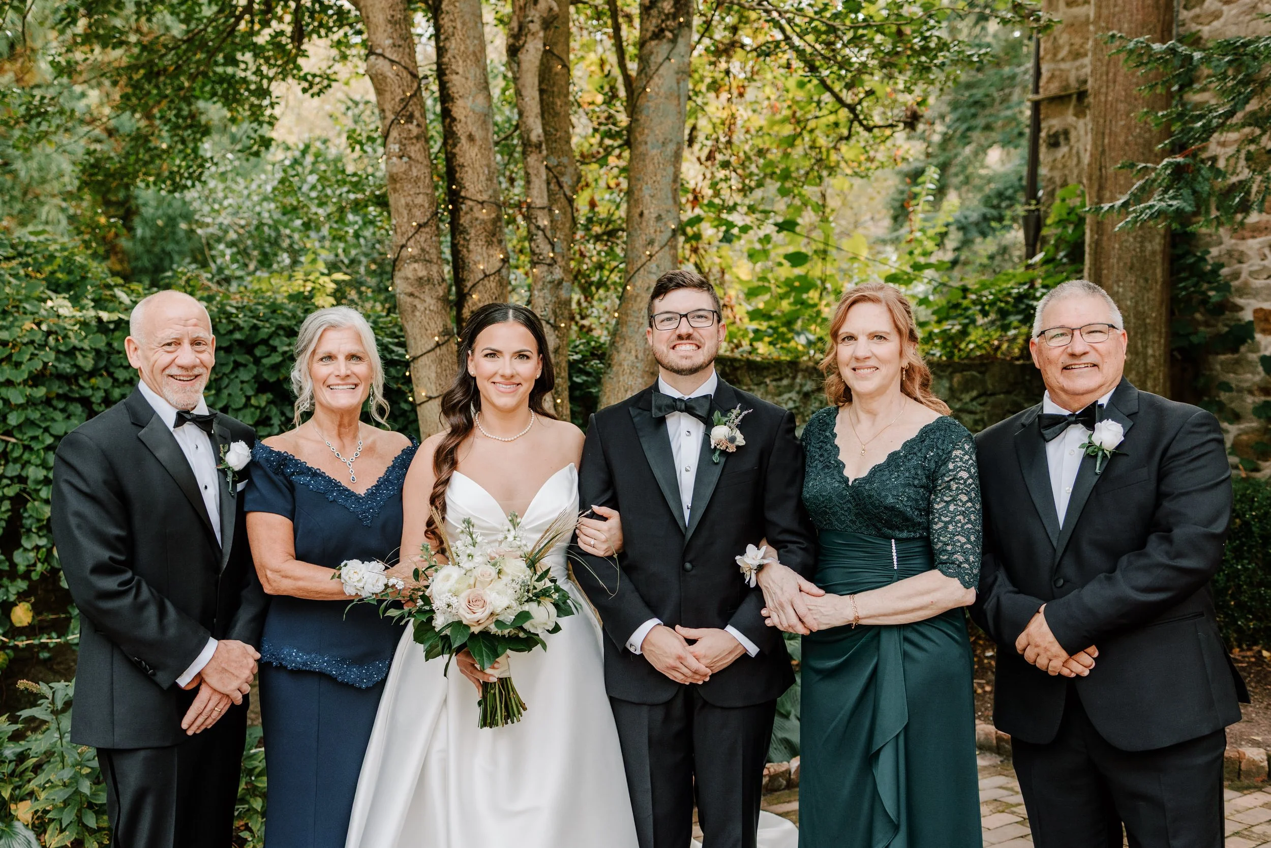 Group of seven people in wedding attire posing outdoors in front of trees and greenery, smiling at the camera.