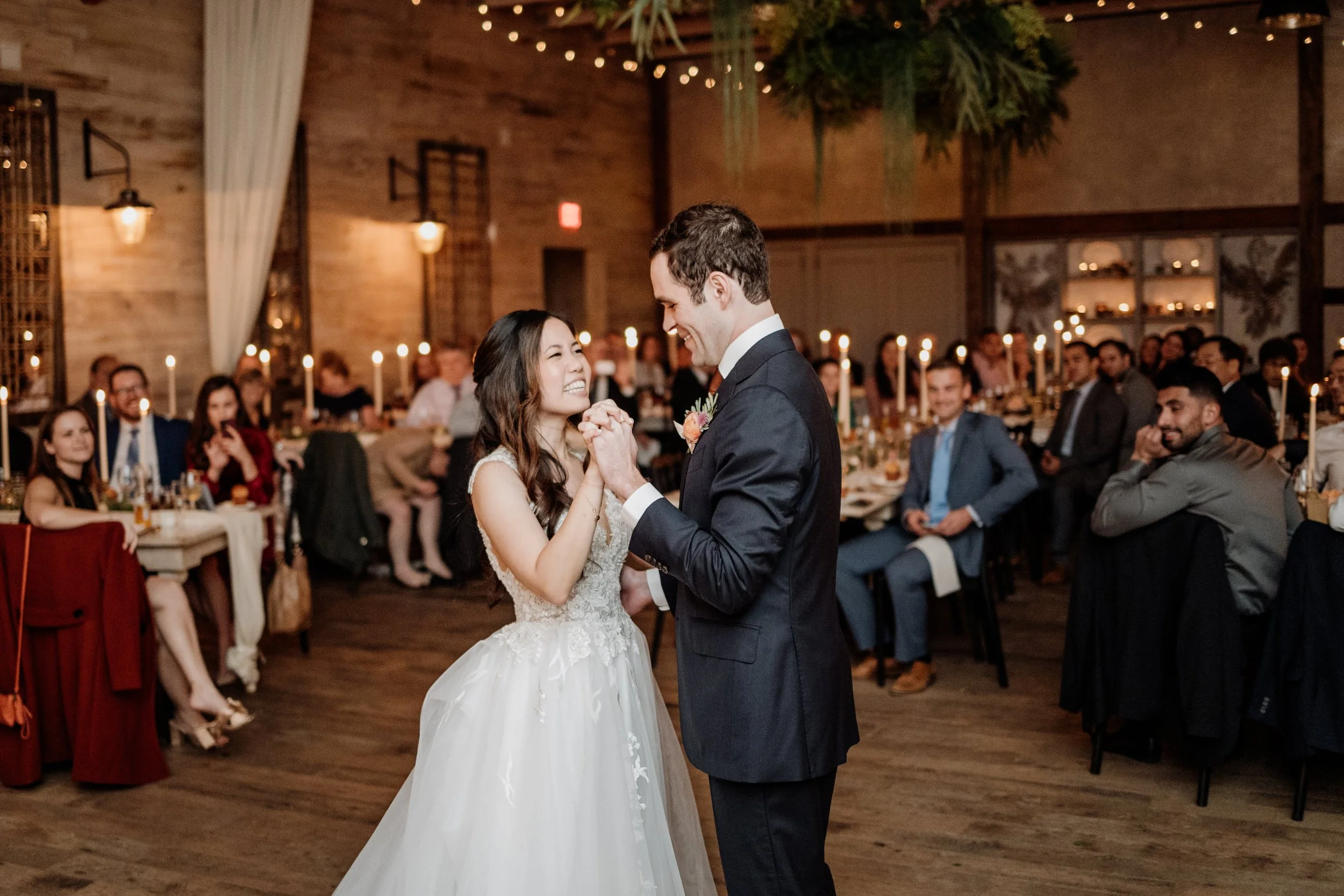 Bride and groom dance at wedding reception with guests seated at tables in the background.