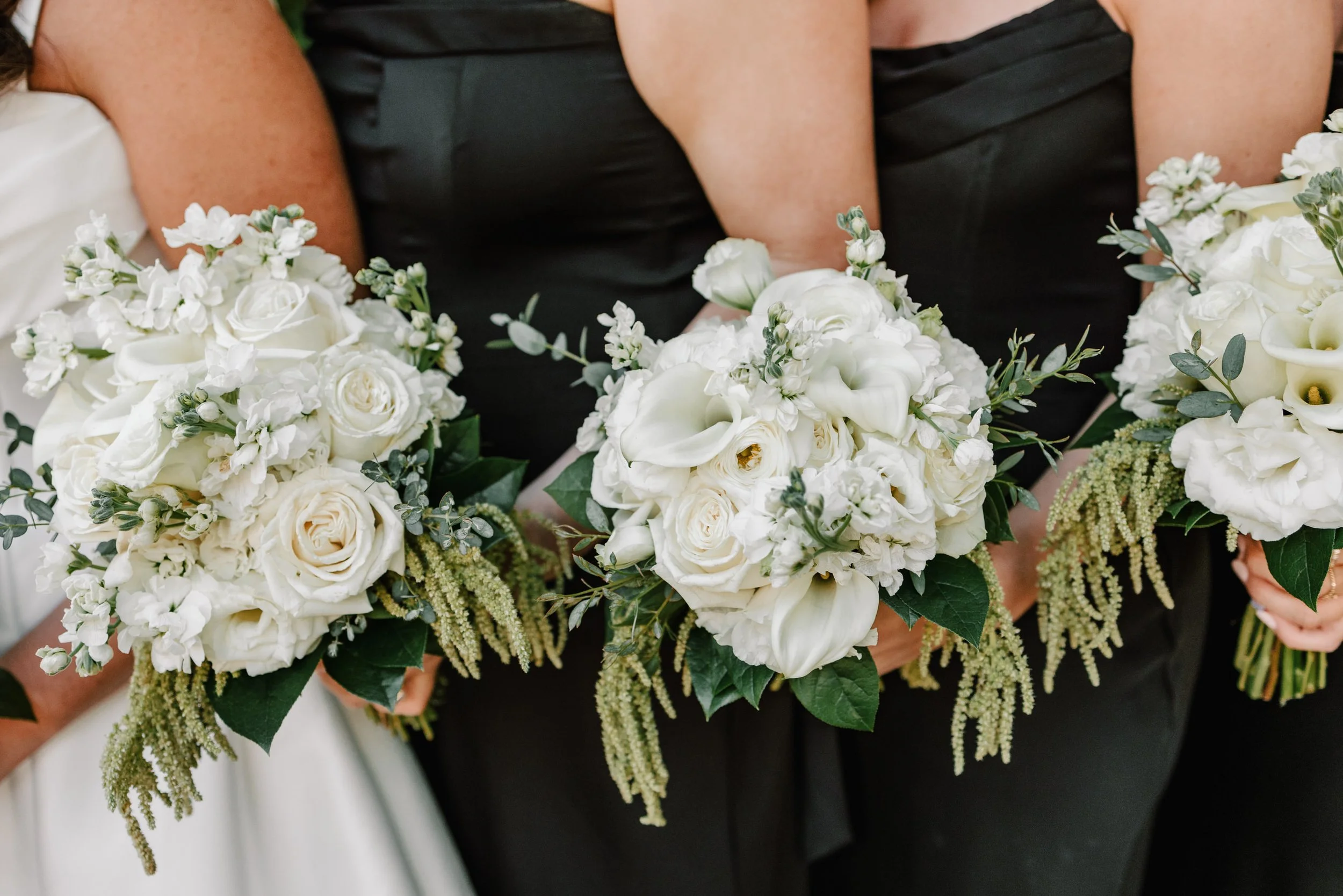 Women holding white floral bouquets at a wedding, with women wearing black dresses.