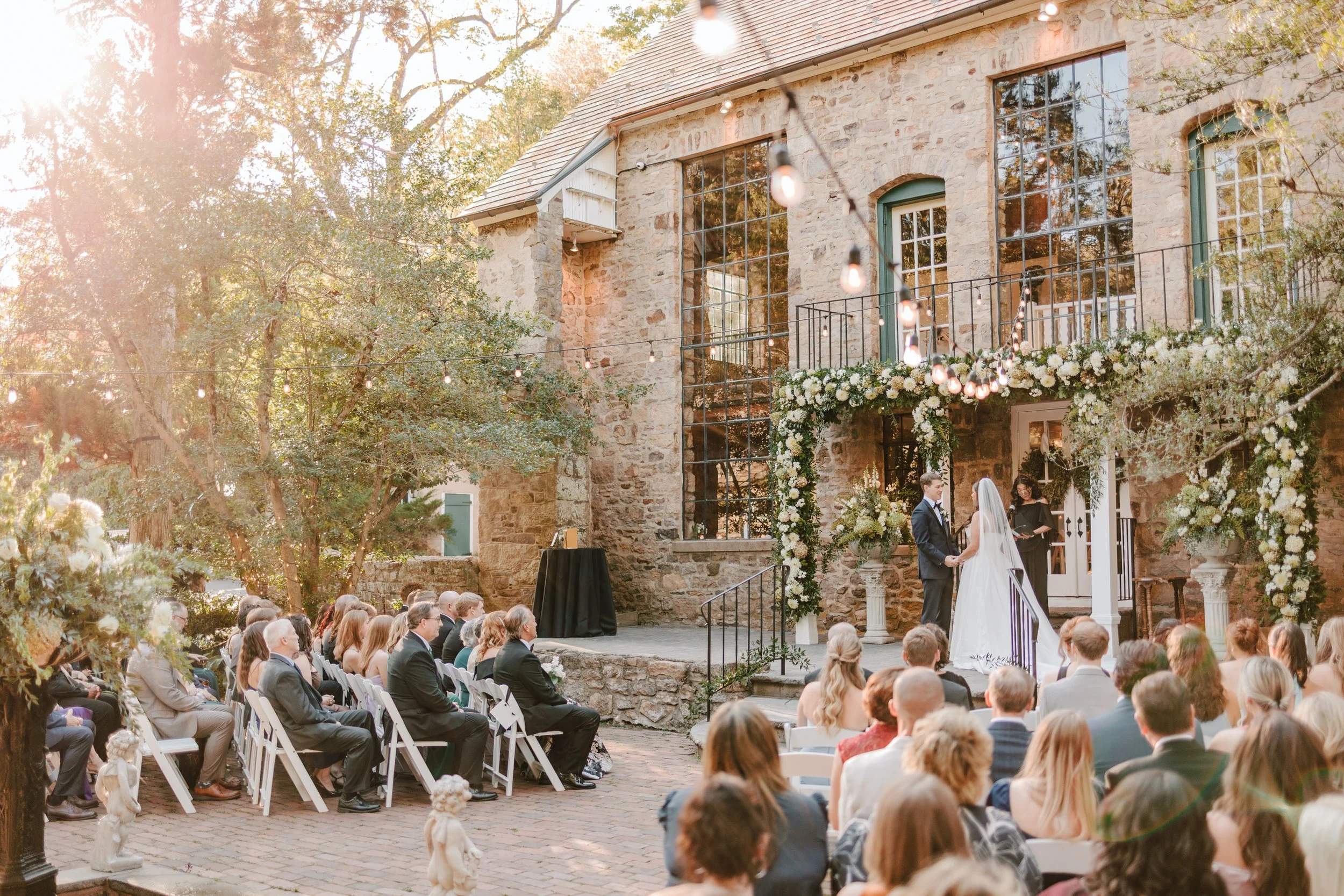 Outdoor wedding ceremony taking place on a brick patio in front of a stone building, with the bride and groom standing under a floral arch, guests seated facing them, and string lights hanging overhead.