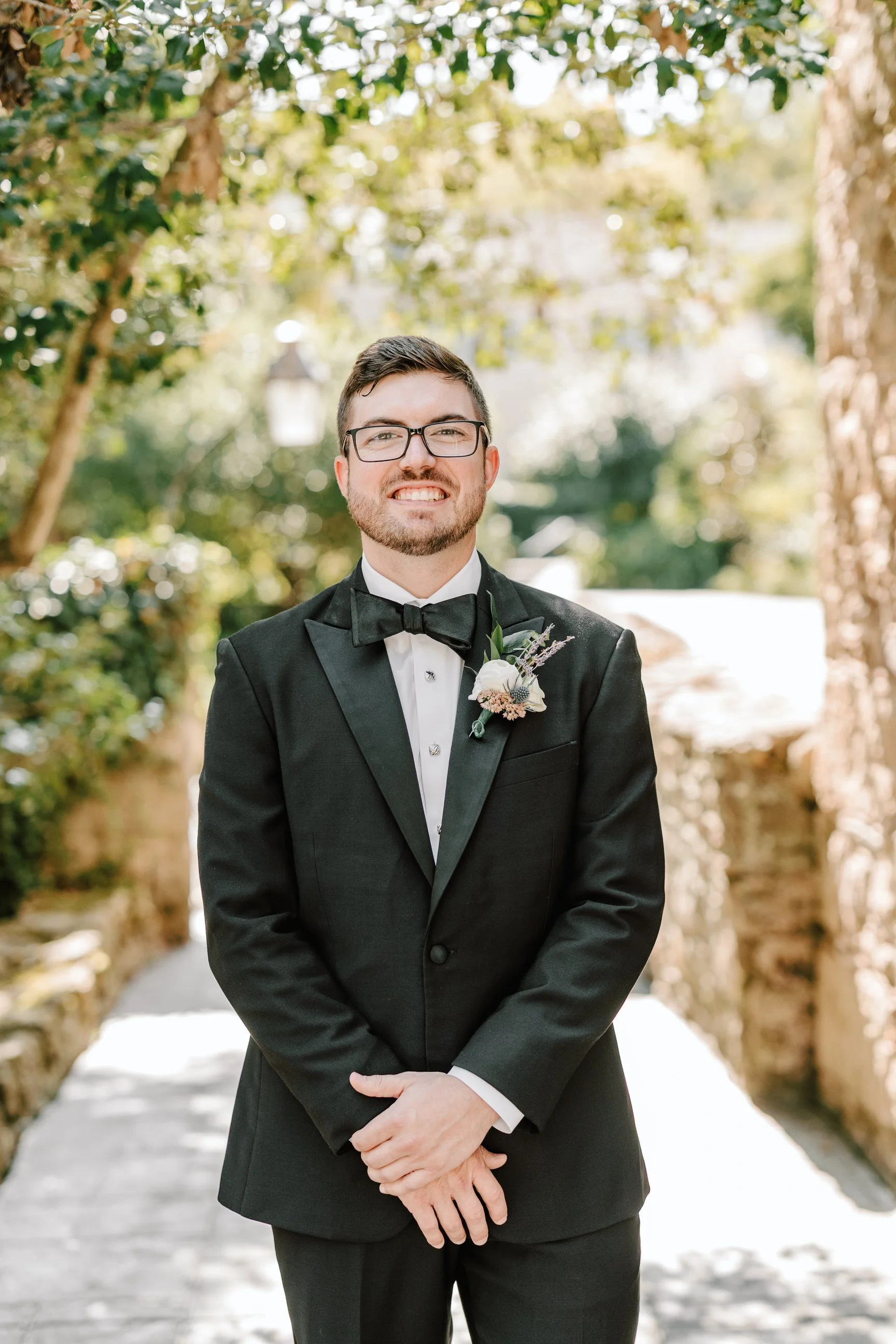A man in a black tuxedo with a bow tie and glasses standing outdoors on a sunny day, smiling, with green trees and a stone wall in the background.