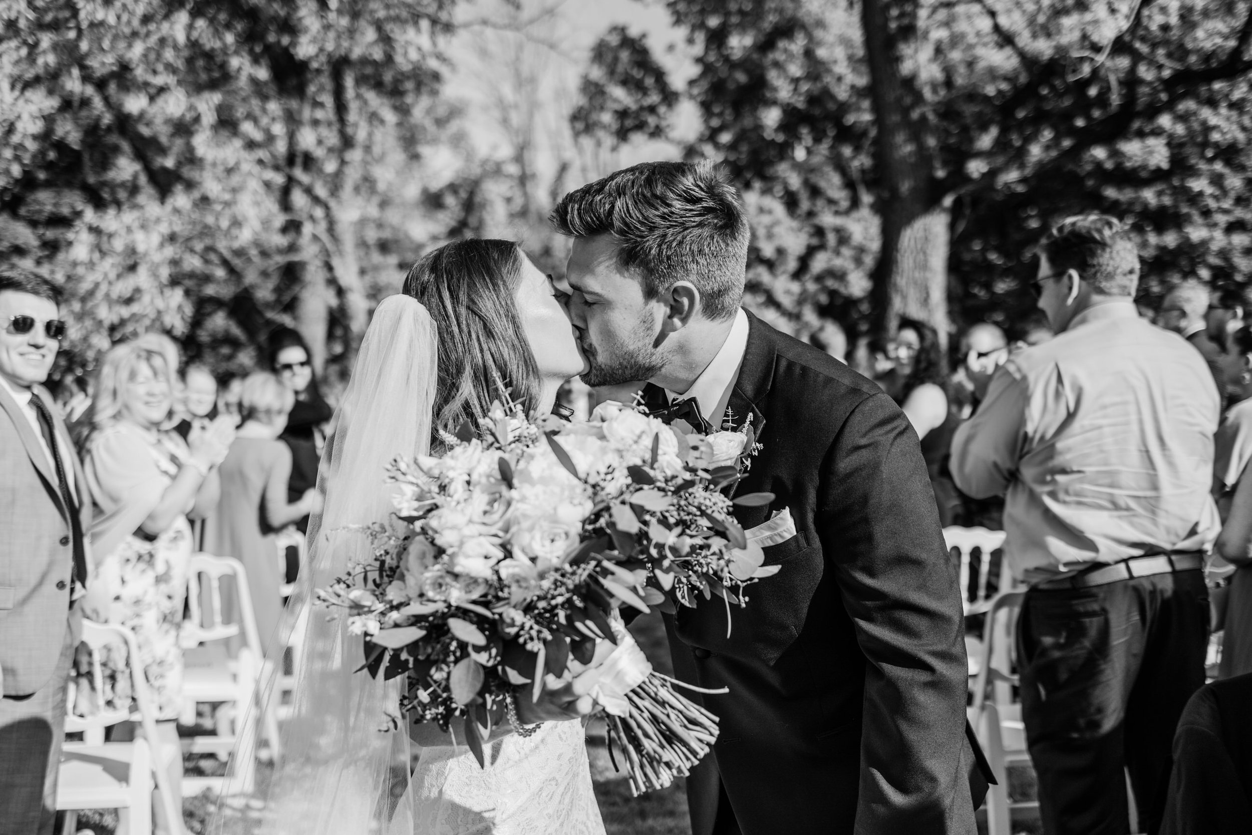A bride and groom sharing a kiss at their outdoor wedding ceremony, surrounded by guests clapping and smiling, with trees in the background.