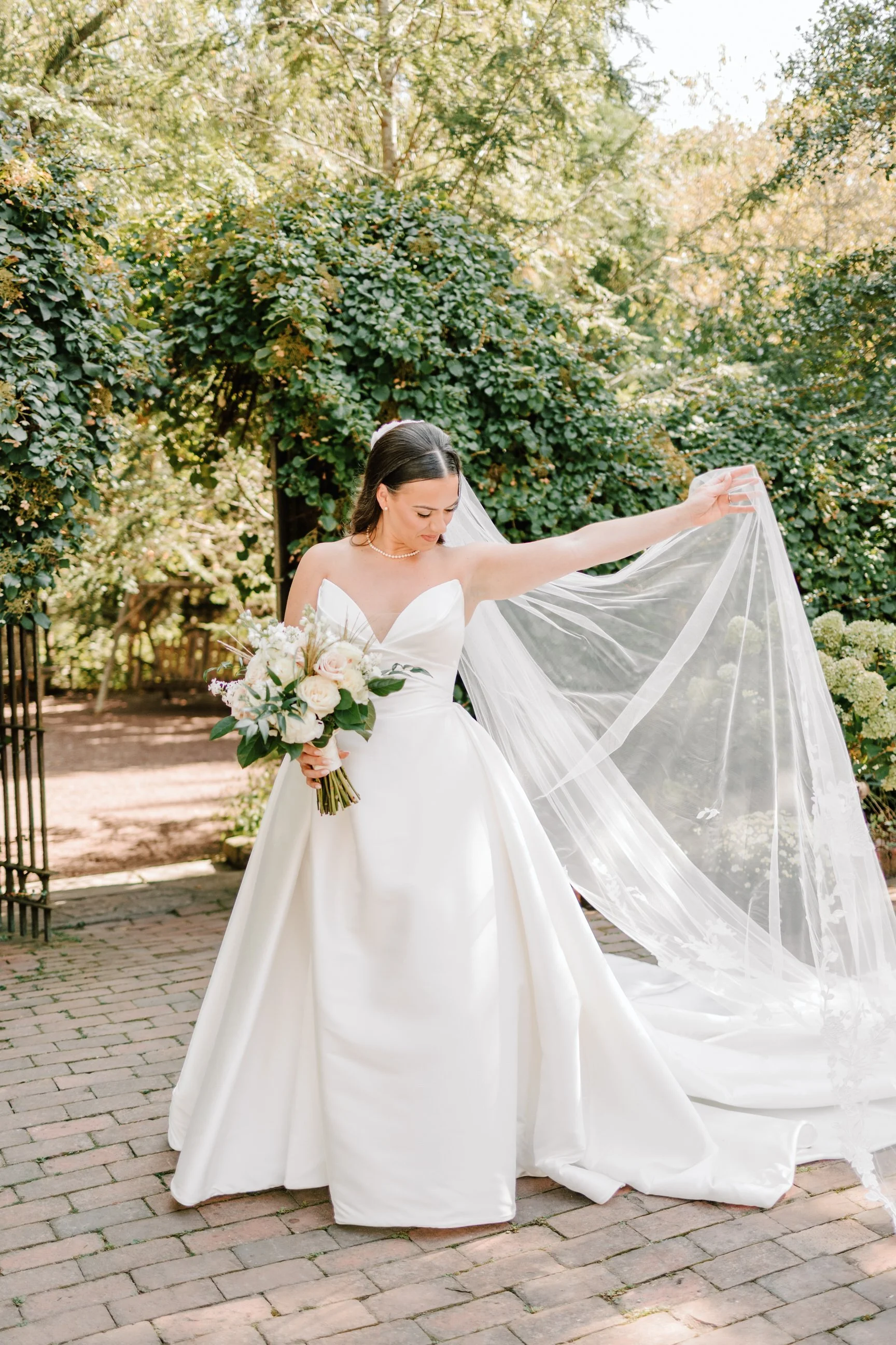 A bride in a white wedding gown holding a bouquet of white flowers, standing outdoors on a brick pathway with green trees in the background, lifting her veil.