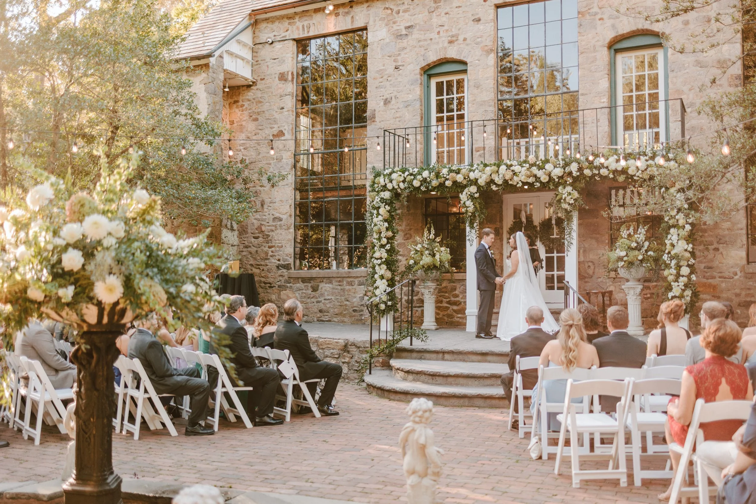 A wedding ceremony taking place outdoors in front of a stone building with large windows. The bride and groom are standing on steps, holding hands, under a floral arch decorated with white flowers and greenery. Guests are seated in white chairs, watc