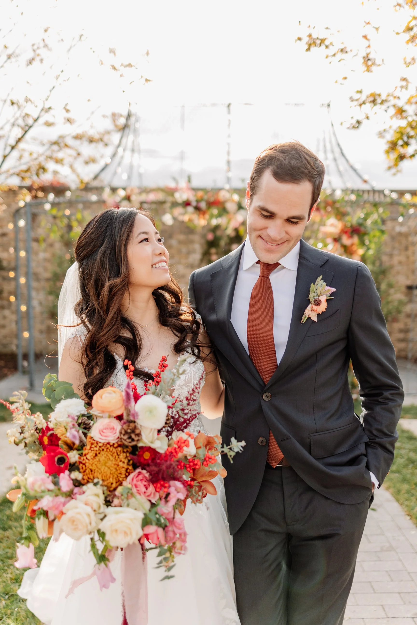 A bride and groom stand close together outdoors, smiling at each other, with a floral bouquet in the bride's hands. The bride has long, wavy brown hair and wears a white lace wedding gown with a veil, while the groom wears a dark suit with a rust-col