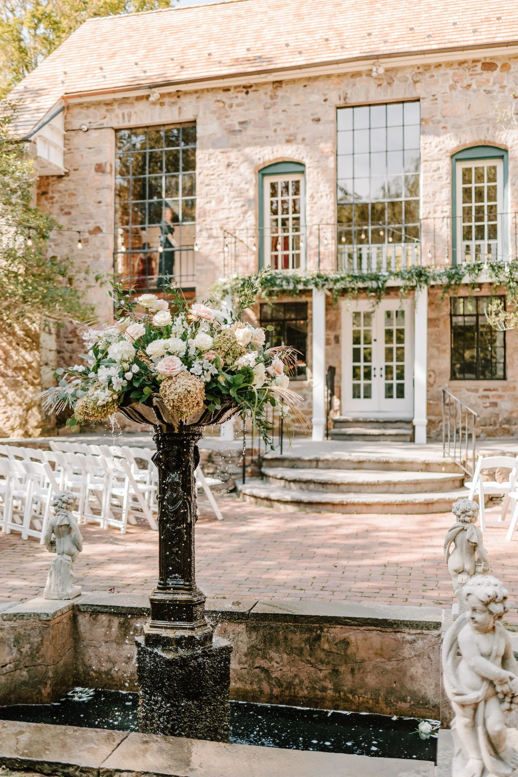 Elegant outdoor wedding setup featuring a stone building with large windows, a fountain with a flower arrangement, and white chairs lining a brick path.