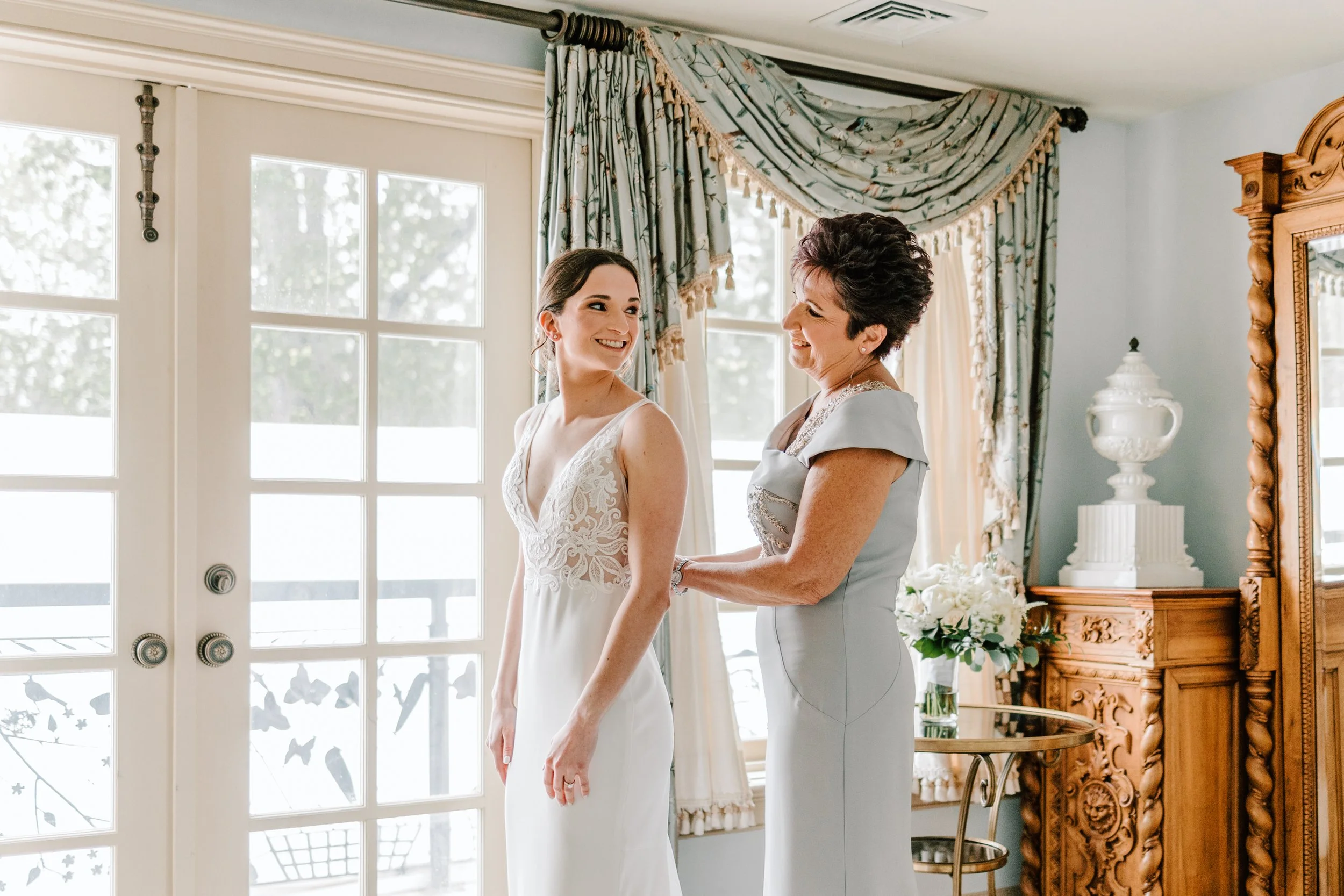 A bride in a white lace wedding dress smiling at an older woman in a light gray dress as she adjusts the back of the bride's dress, in a room with large windows and floral curtains.