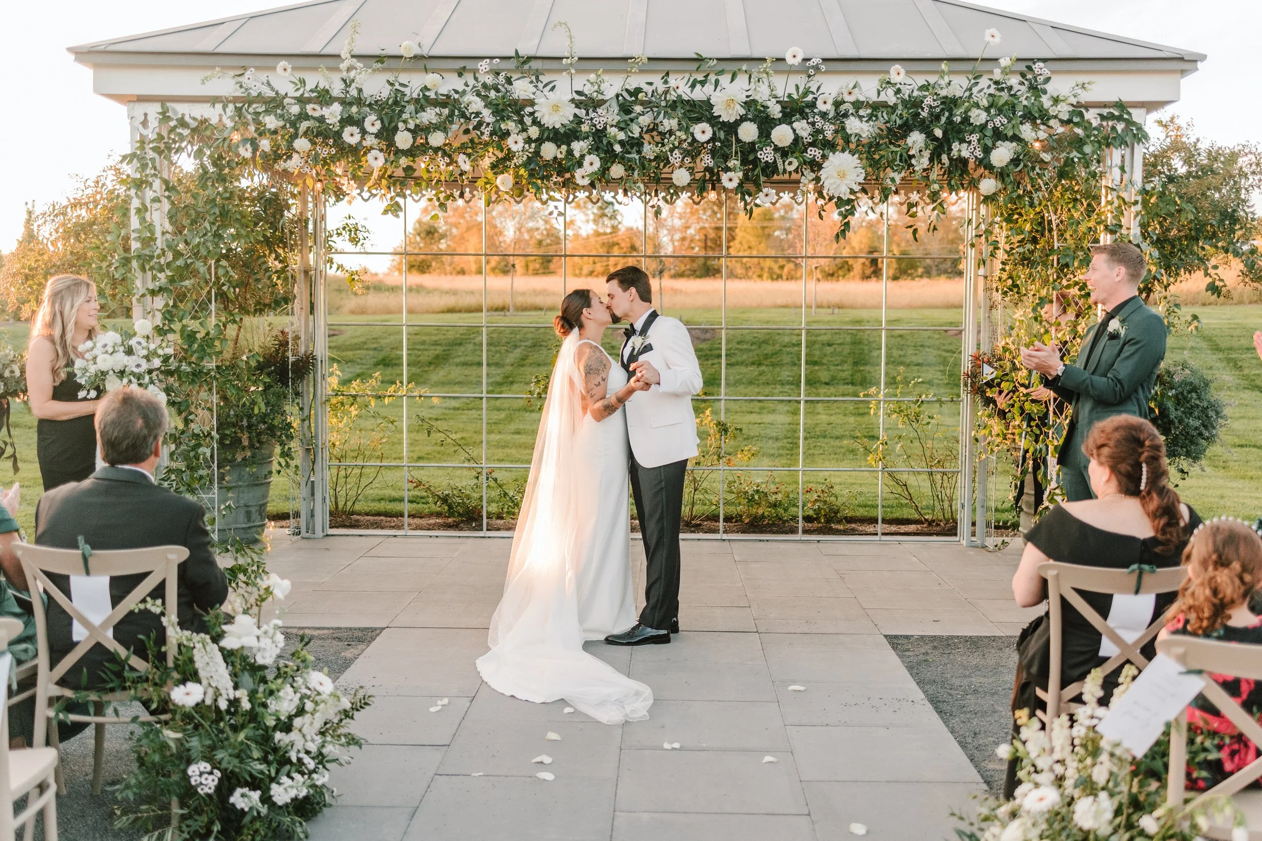 Wedding ceremony with couple kissing under a flower-decorated gazebo, surrounded by guests seated and standing, outdoors in a field setting.