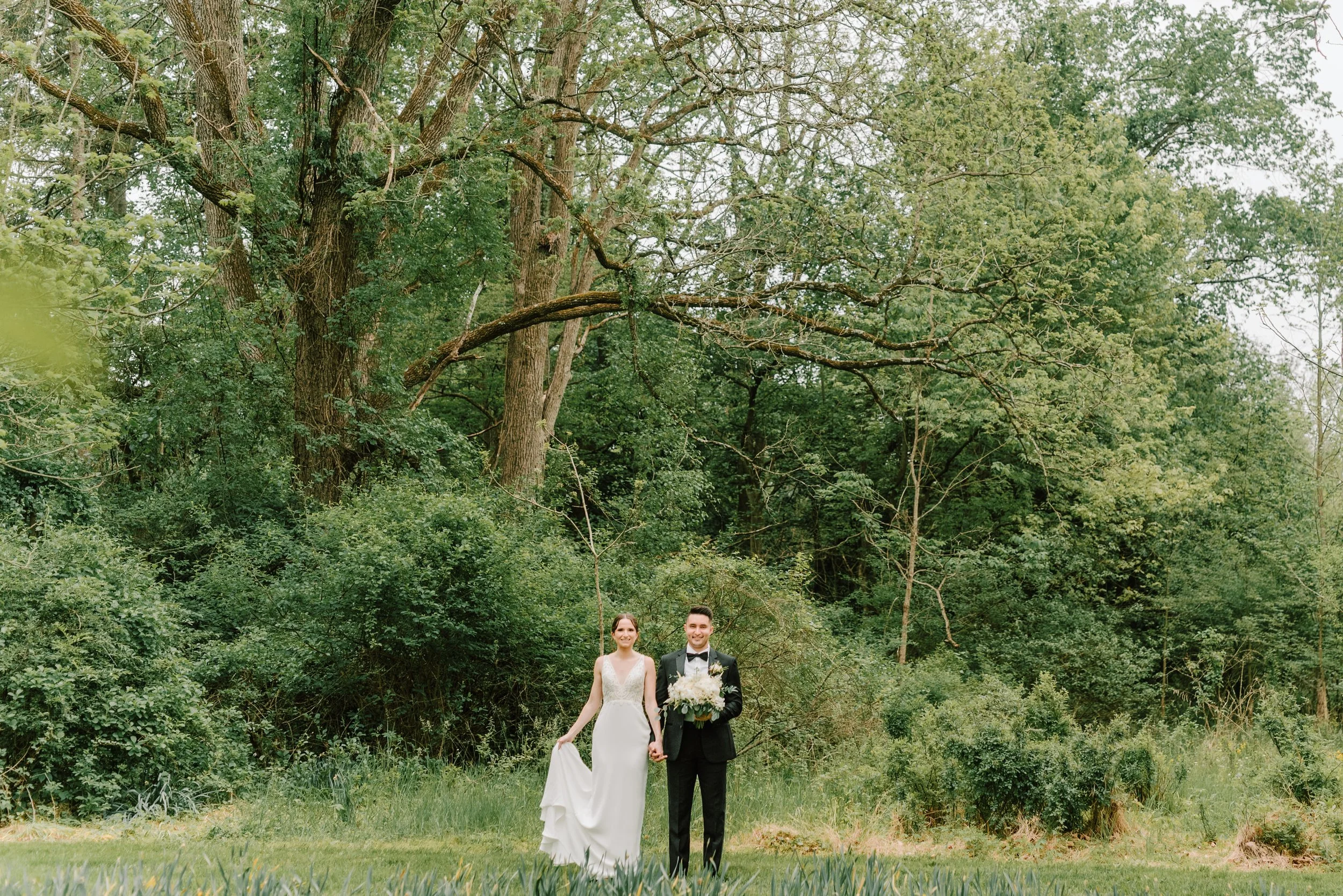 A bride and groom walking hand in hand outdoors in a lush, green forest setting. The bride is dressed in a white wedding gown, and the groom is wearing a black tuxedo with a bow tie and holding a bouquet of white flowers. They are smiling and looking