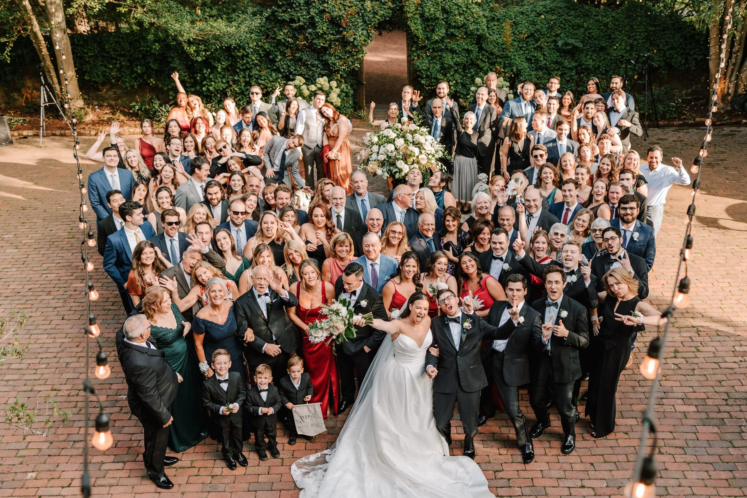 A large wedding celebration with the bride and groom in the front, surrounded by guests on a brick-paved outdoor area, with string lights overhead and greenery in the background.