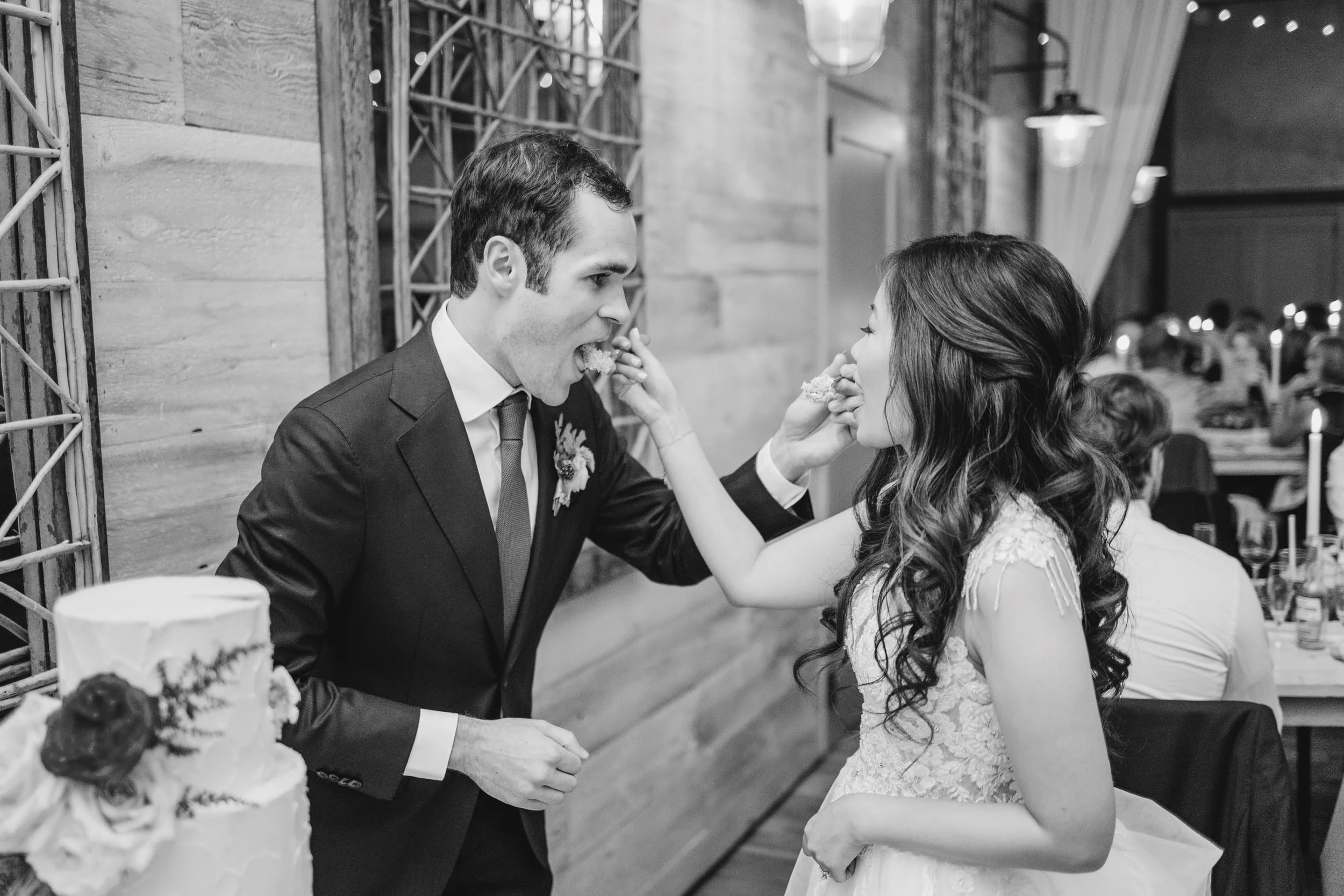 A couple at a wedding reception feeding each other cake. The groom is wearing a suit and tie, and the bride has long wavy hair and a lace dress. Guests are seated at tables in the background.