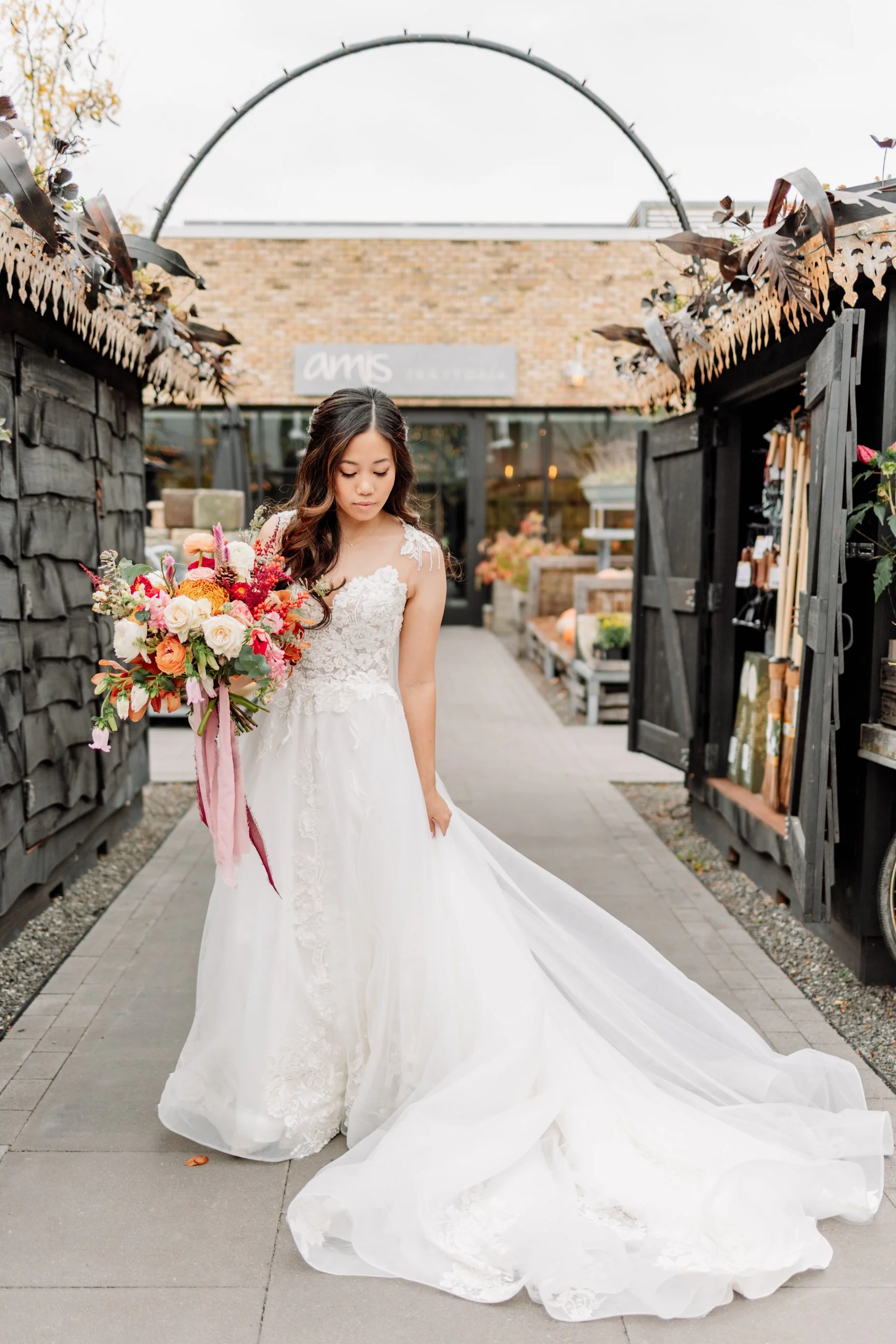 Bride in white wedding gown holding a colorful bouquet, walking outdoors near a black shingled fence and open door, with a brick building in the background.