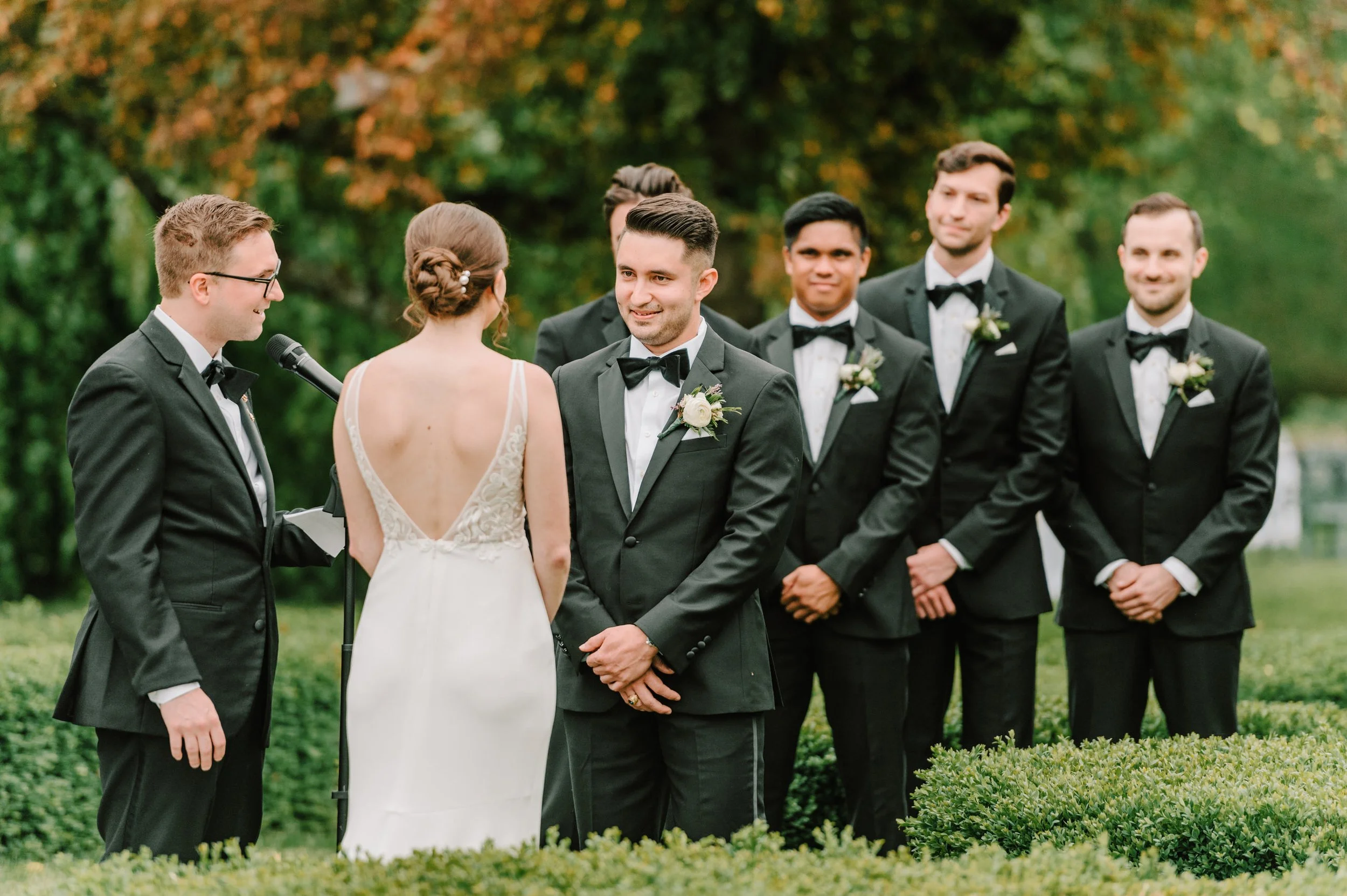 A wedding ceremony outdoors with the bride and groom exchanging vows, surrounded by the bridal party dressed in black tuxedos, under trees with autumn foliage.