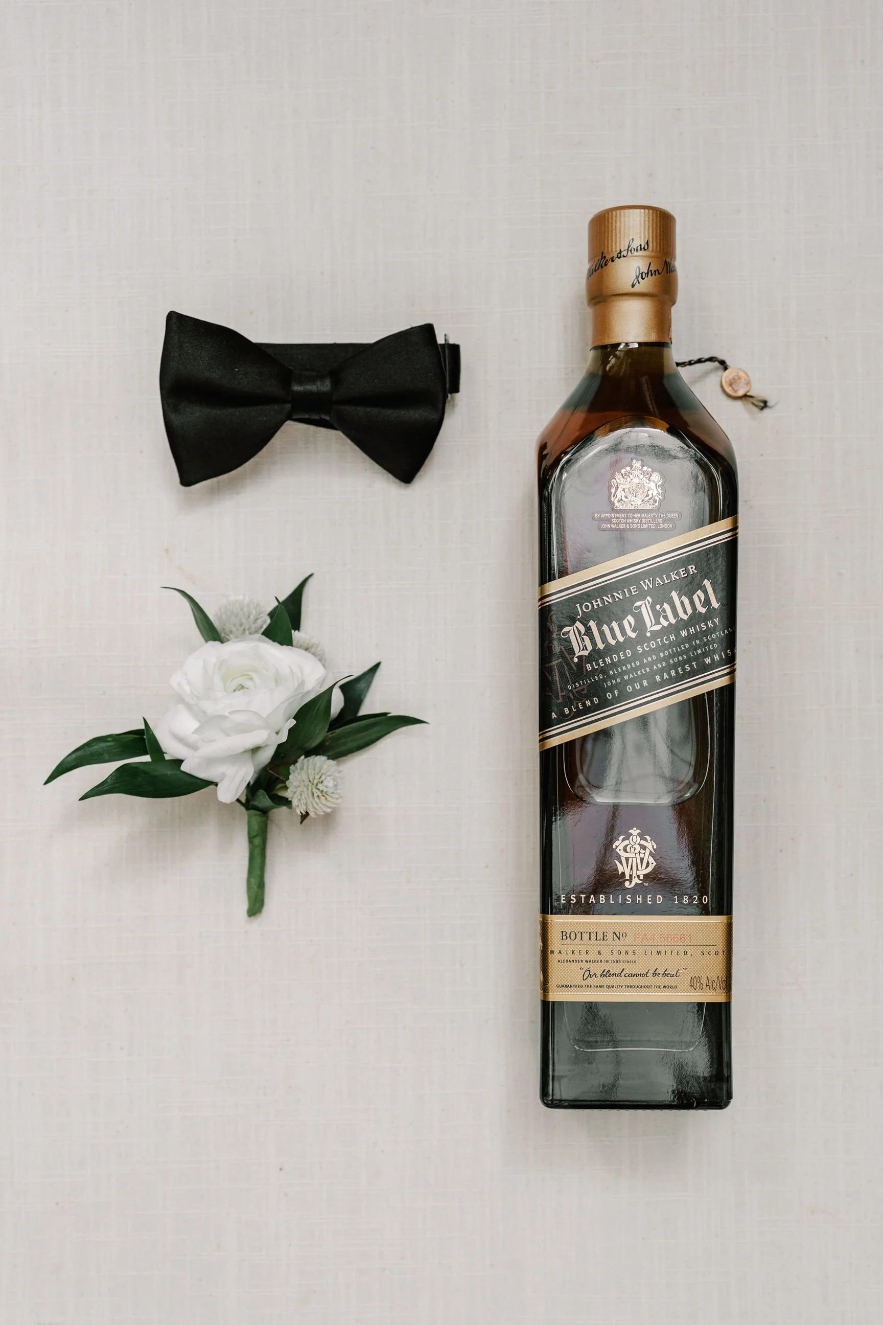 A black bow tie, a white boutonniere with green leaves, and a bottle of Johnnie Walker Blue Label whisky on a white background.