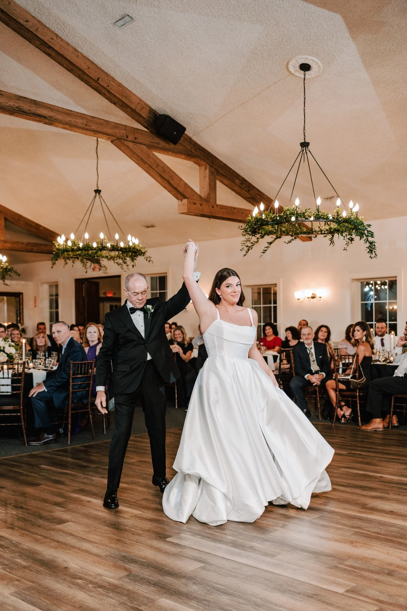 A bride and an older man, likely her father, dancing at a wedding reception, with guests seated at tables in the background.