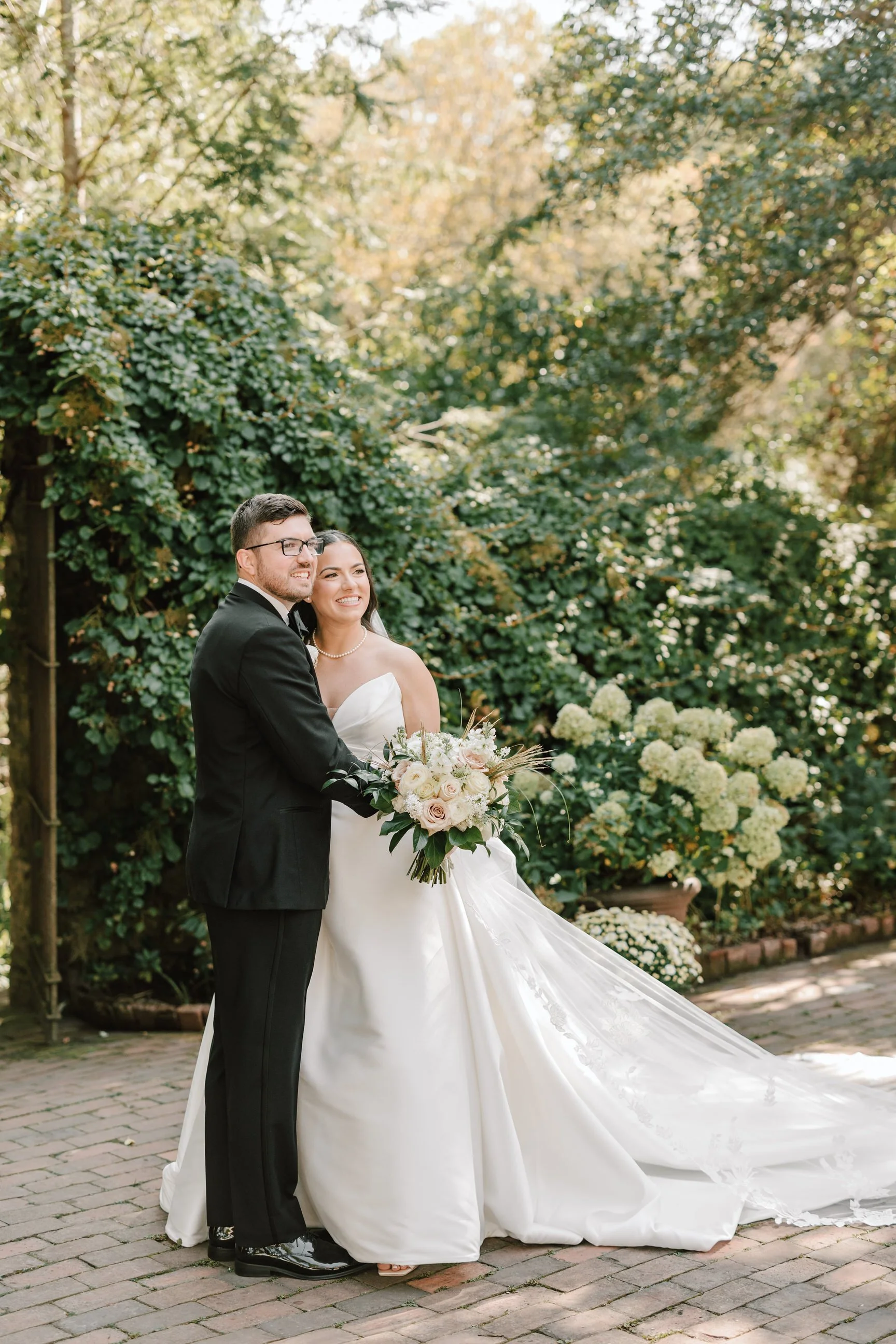Bride and groom in wedding attire standing outdoors in a garden with lush greenery, holding a bouquet of white and blush roses, smiling together.