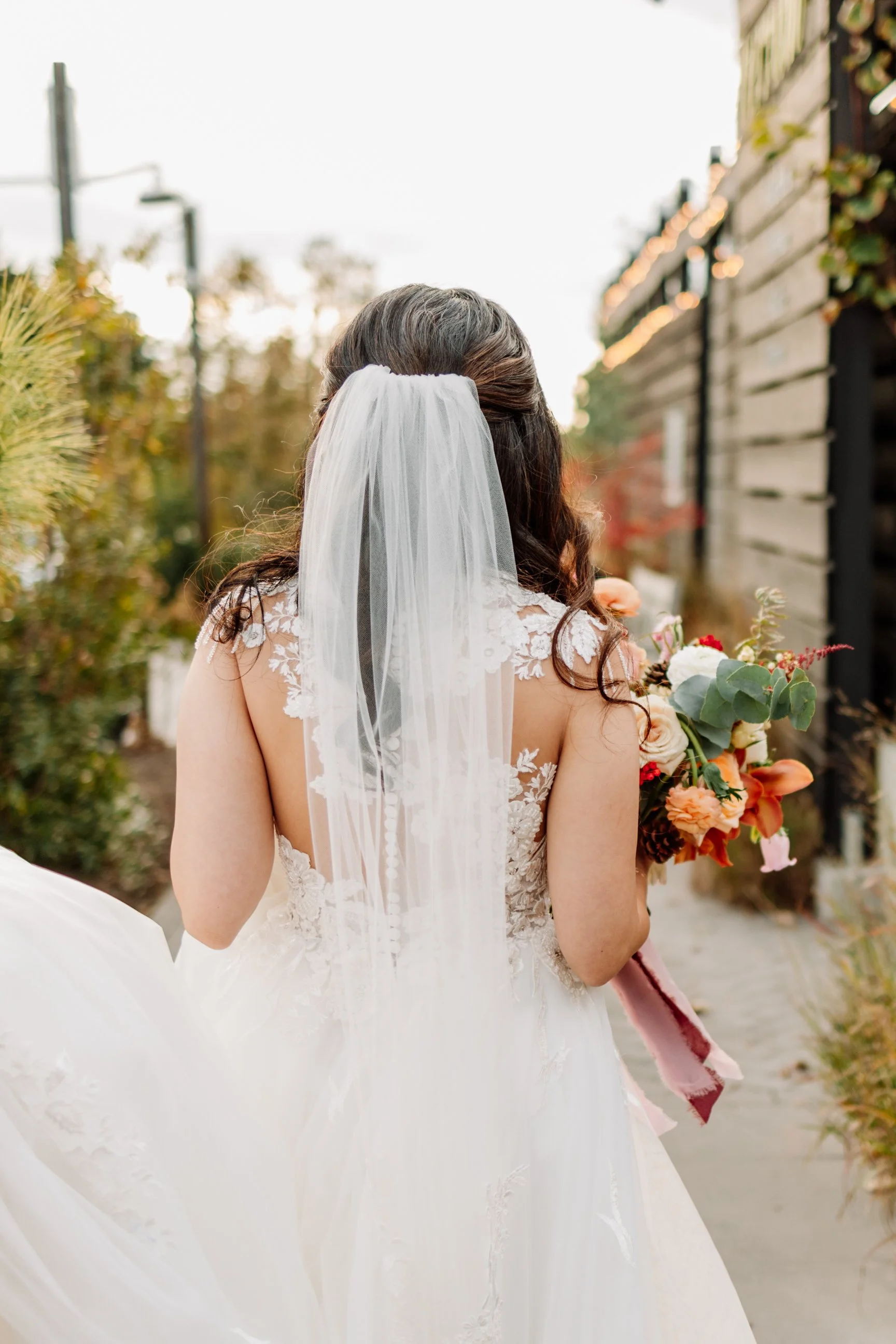 Back view of a bride in a wedding dress holding a bouquet outdoors during sunset.